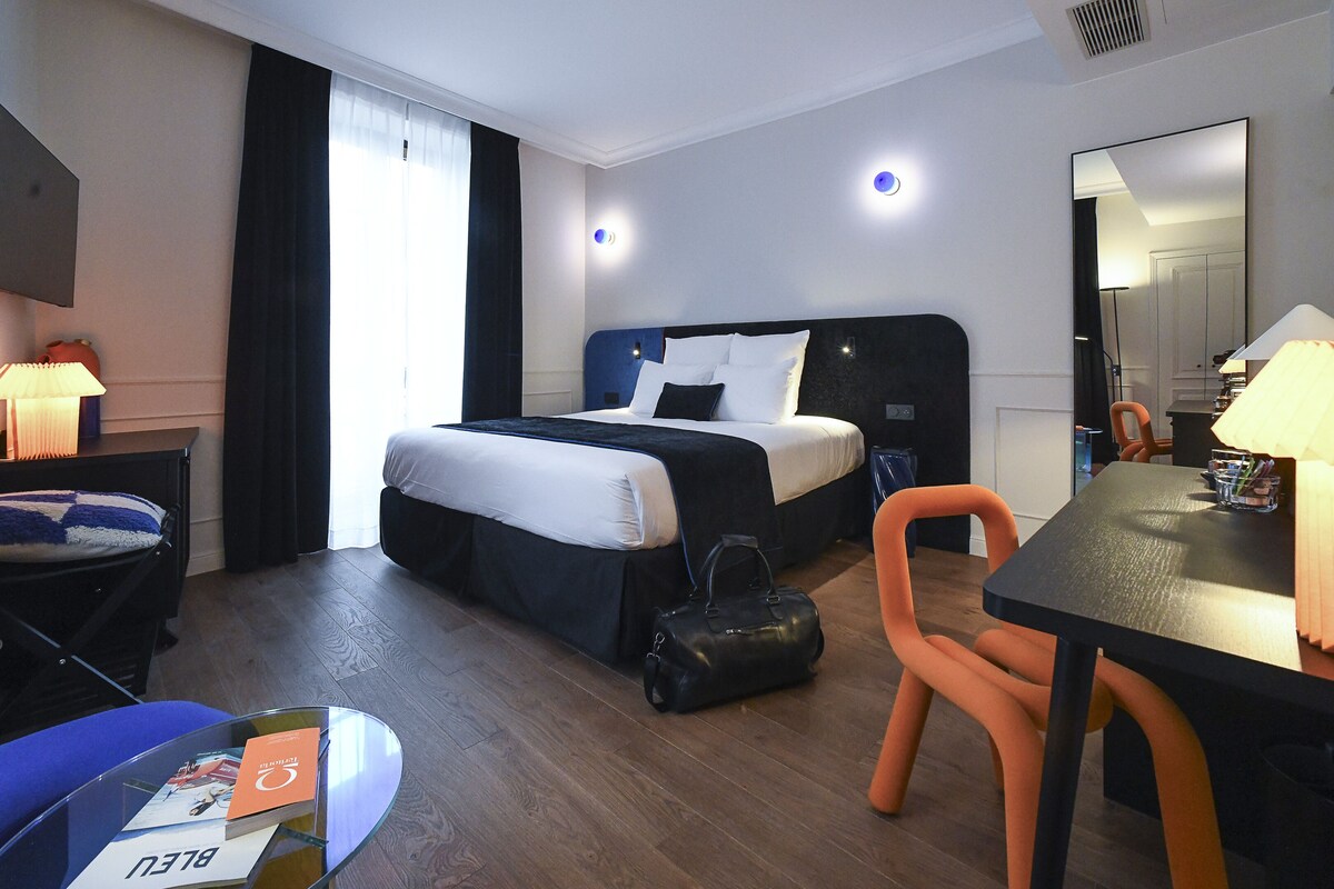 Een hotelkamer met een bed, een bureau met stoel, een tafel met lamp, een handtas en een spiegel.