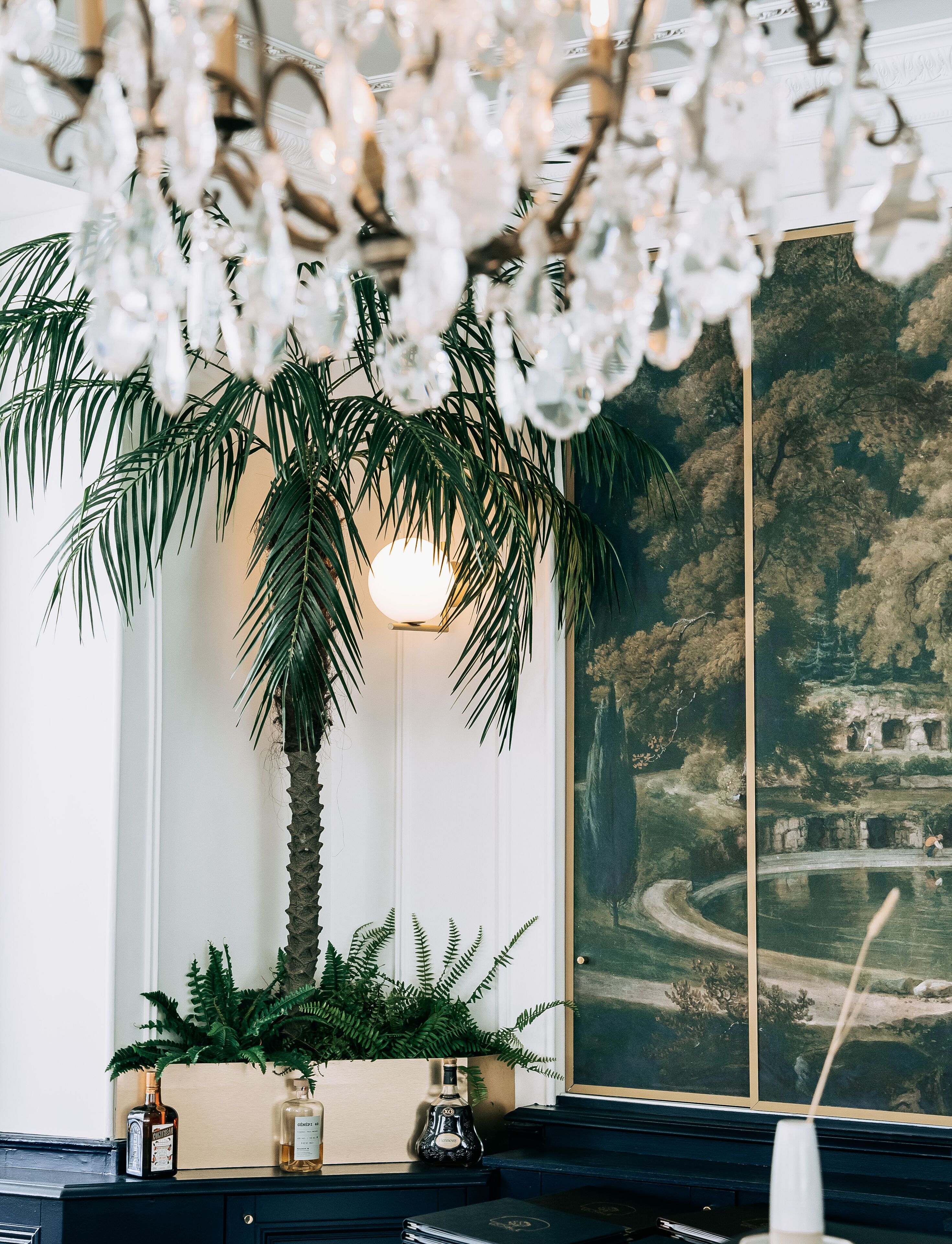 A room with a large painting of a landscape, a palm tree, and a table with bottles and a candle.