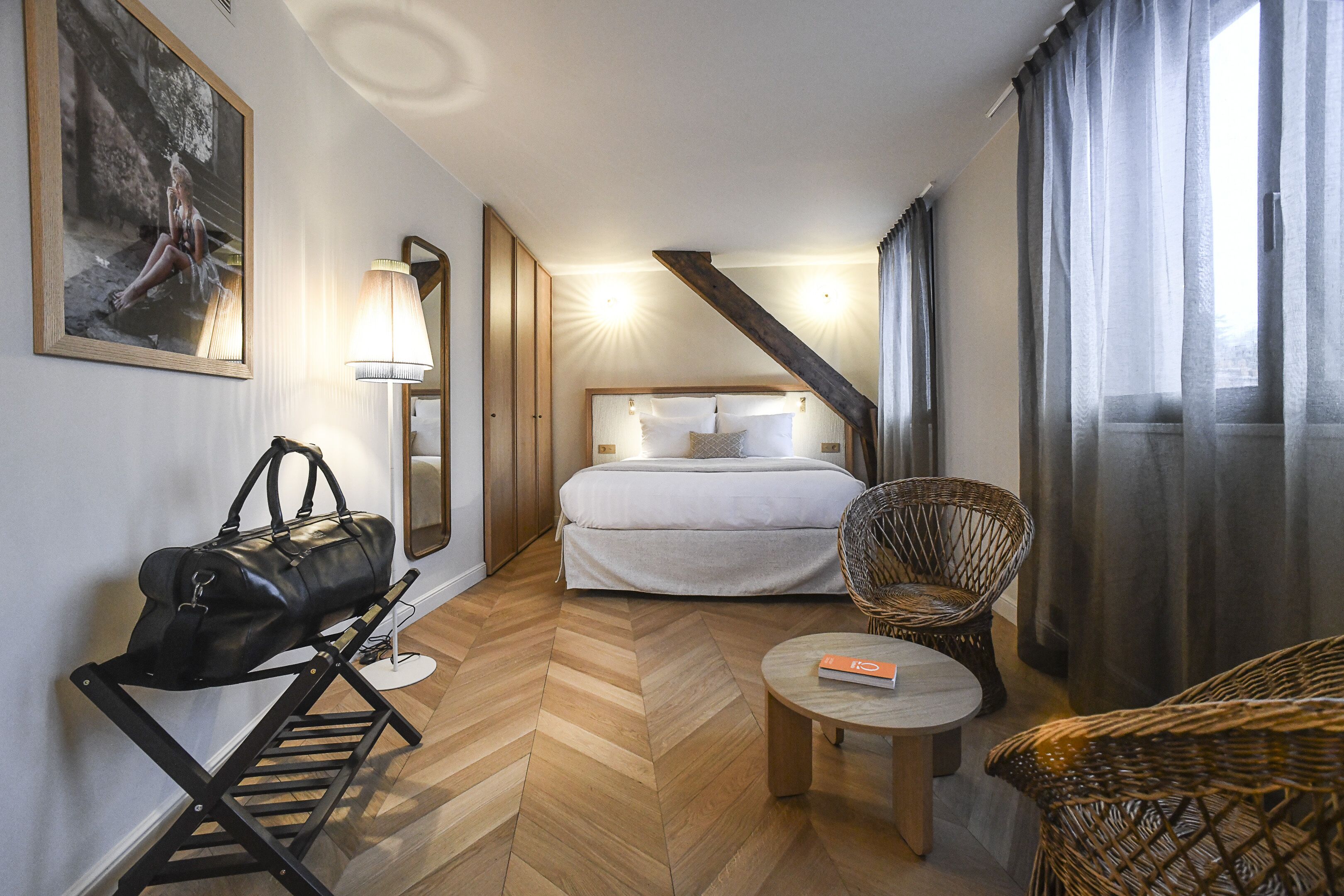 Ein Hotelzimmer mit Bett, Sessel, kleinem Tisch, Lampe und einer Handtasche auf einem Ständer.