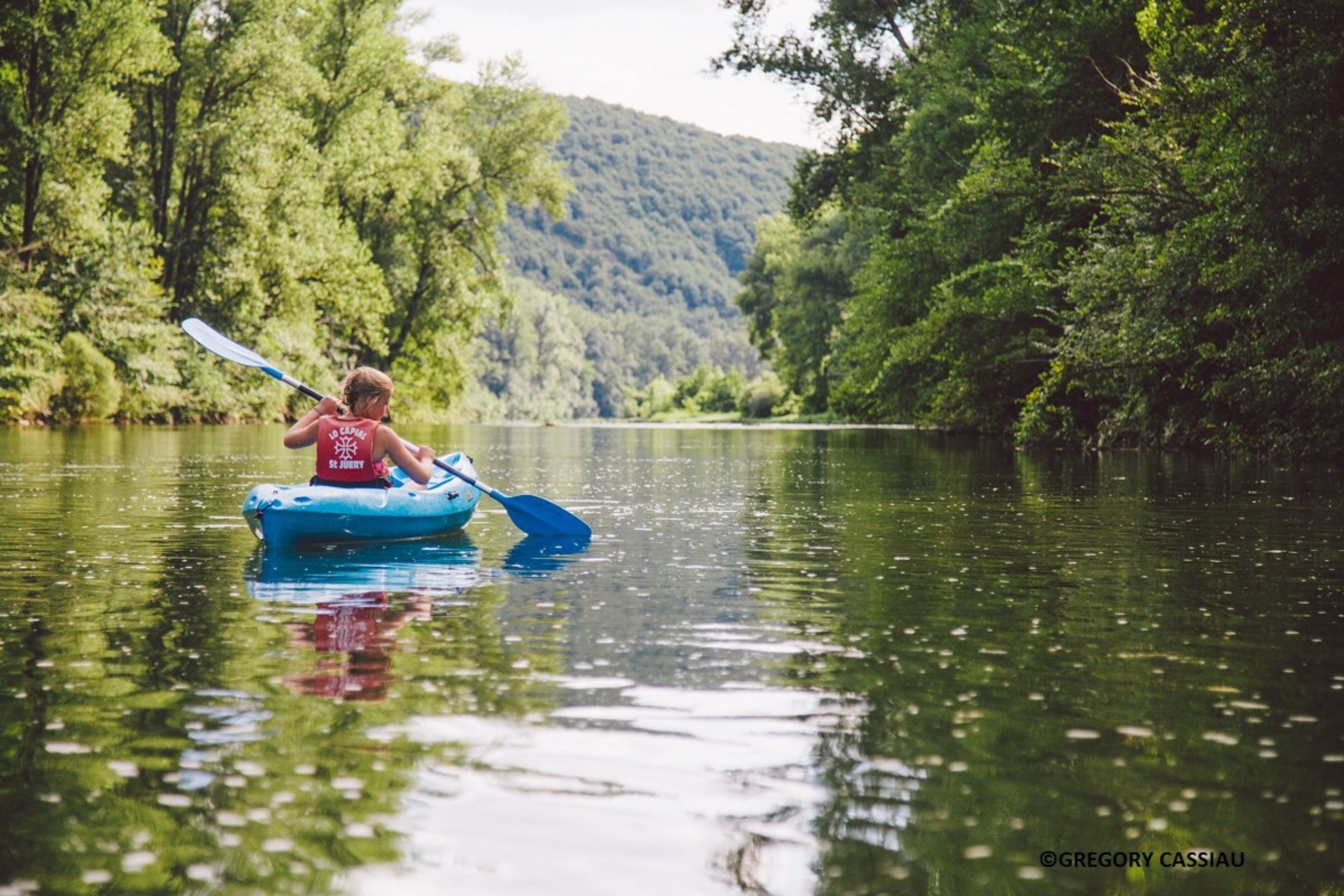 Una persona practica kayak en un río tranquilo rodeado de exuberante vegetación y colinas.