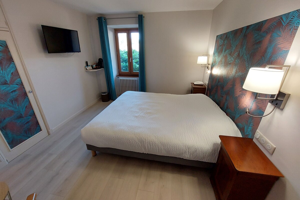 A bedroom with a bed, a TV mounted on the wall, a bedside table with a lamp, and a window with curtains.