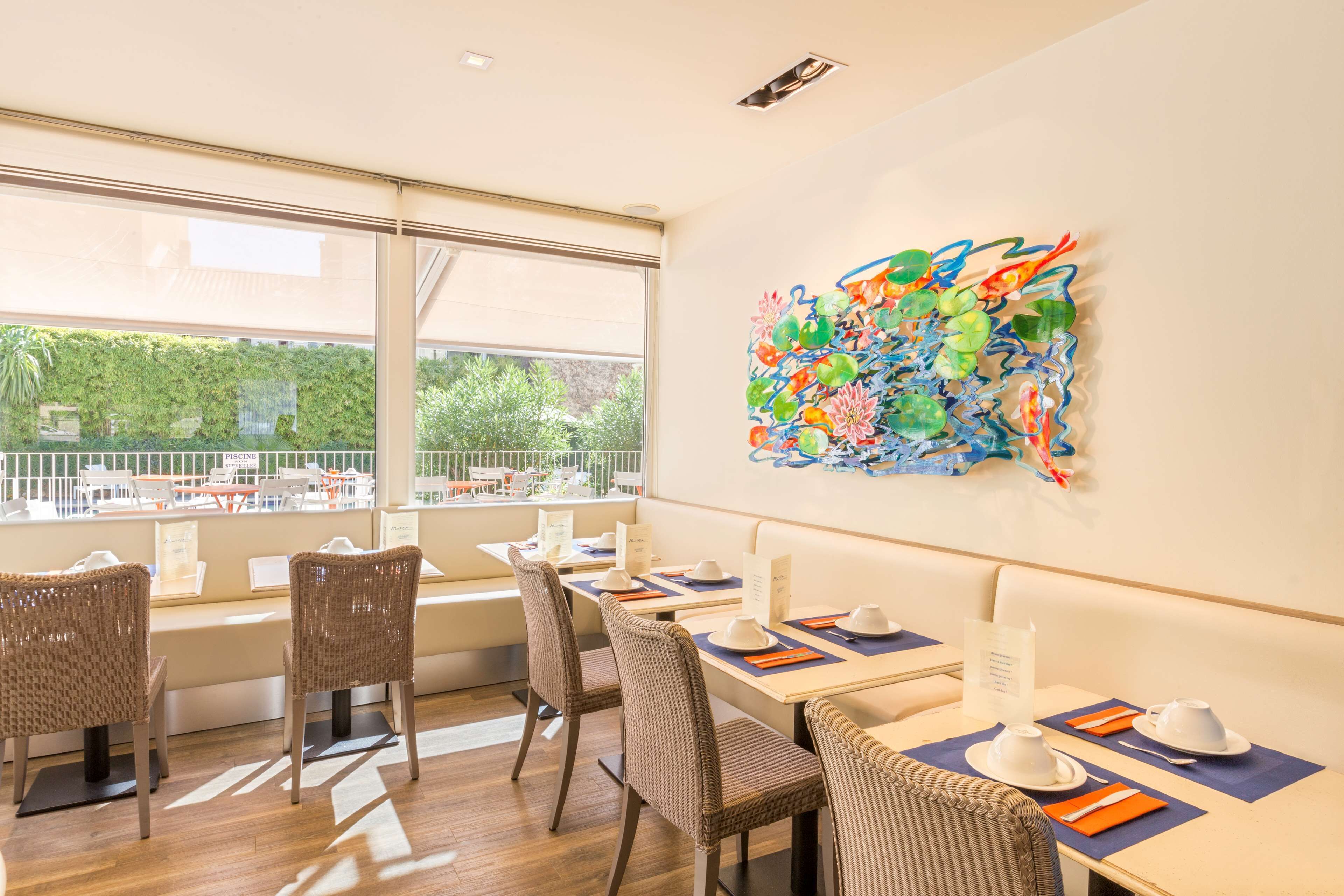 Un comedor con sillas de mimbre, mesas dispuestas con platos blancos y un cuadro abstracto de colores en la pared.