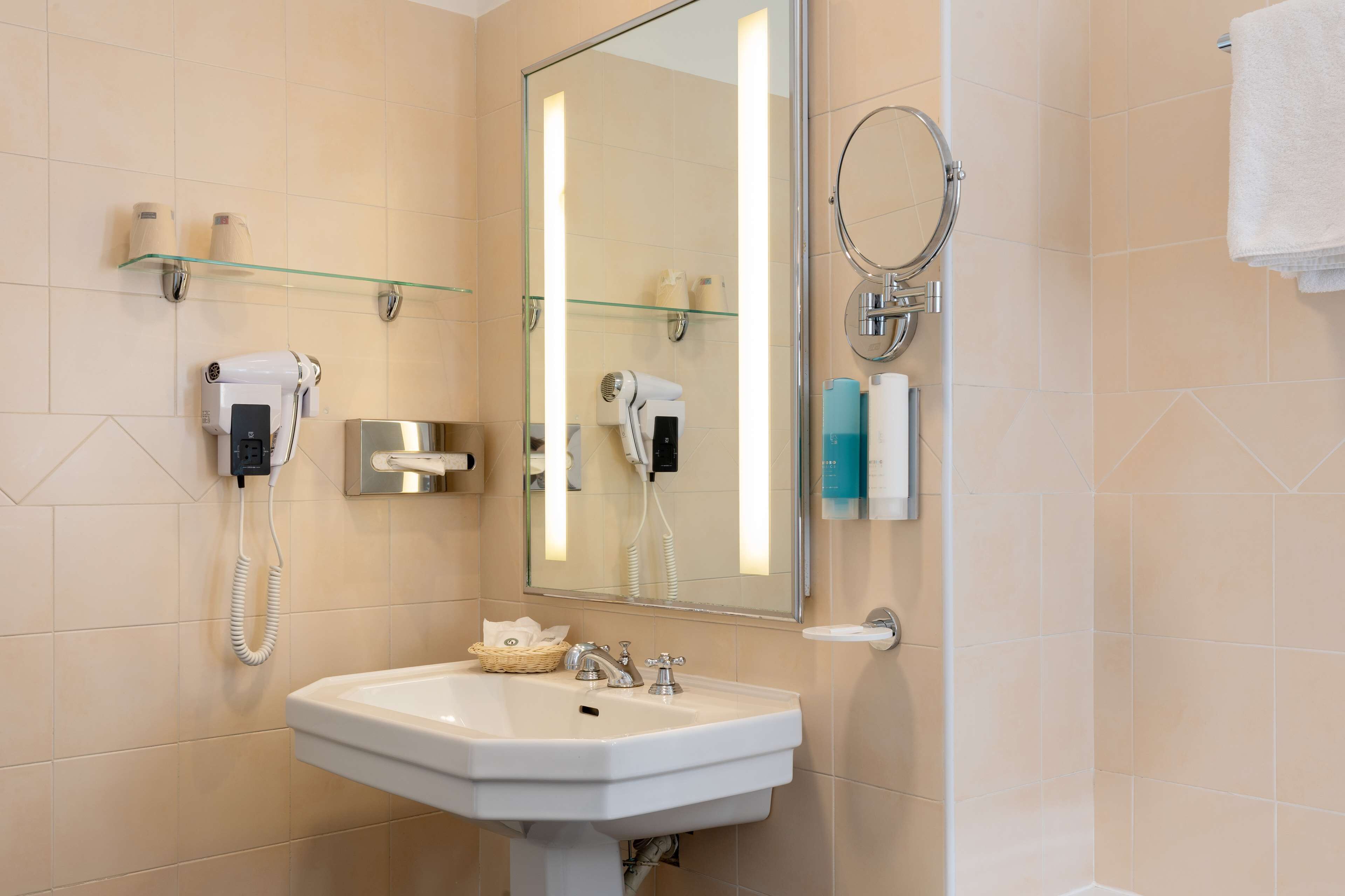 A bathroom with a white sink, a glass shelf with toiletries, a hairdryer, and a mirror.