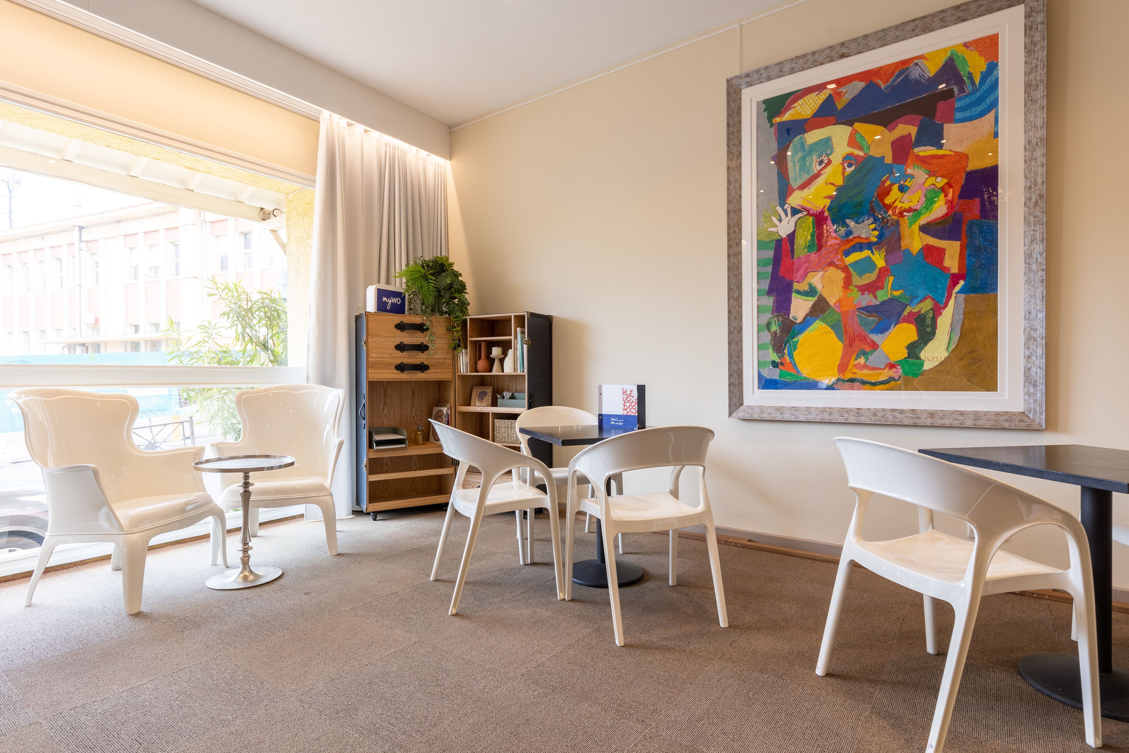 A modern hotel room with a large window, a dining area with white chairs and a small table, a bookshelf, and a colorful abstract painting on the wall.