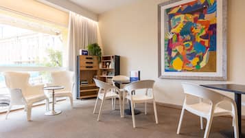 A modern hotel room with a large window, a dining area with white chairs and a small table, a bookshelf, and a colorful abstract painting on the wall.