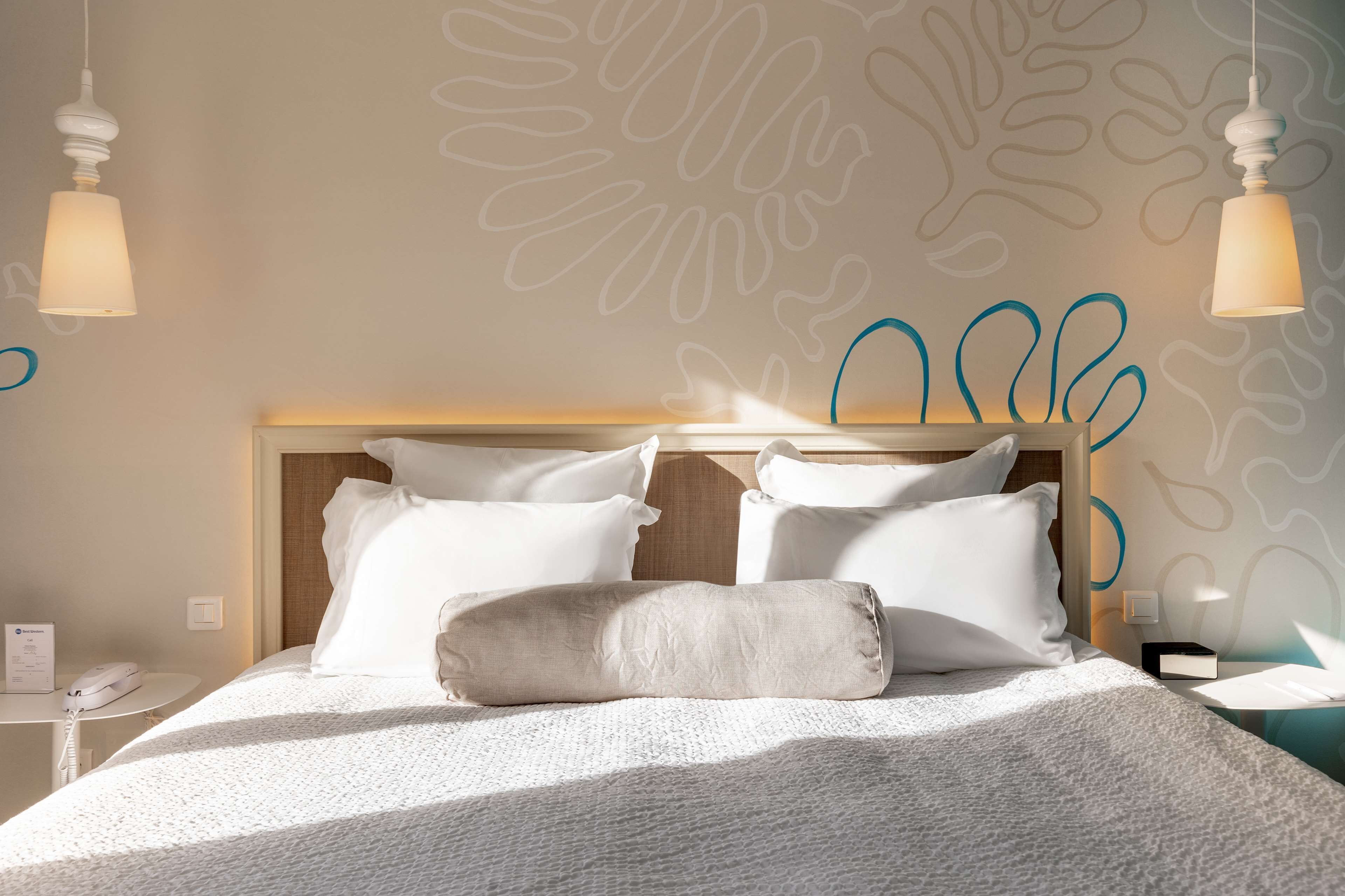 A neatly made bed with white linens and pillows, a wooden headboard, and a wall with abstract blue and beige patterns.