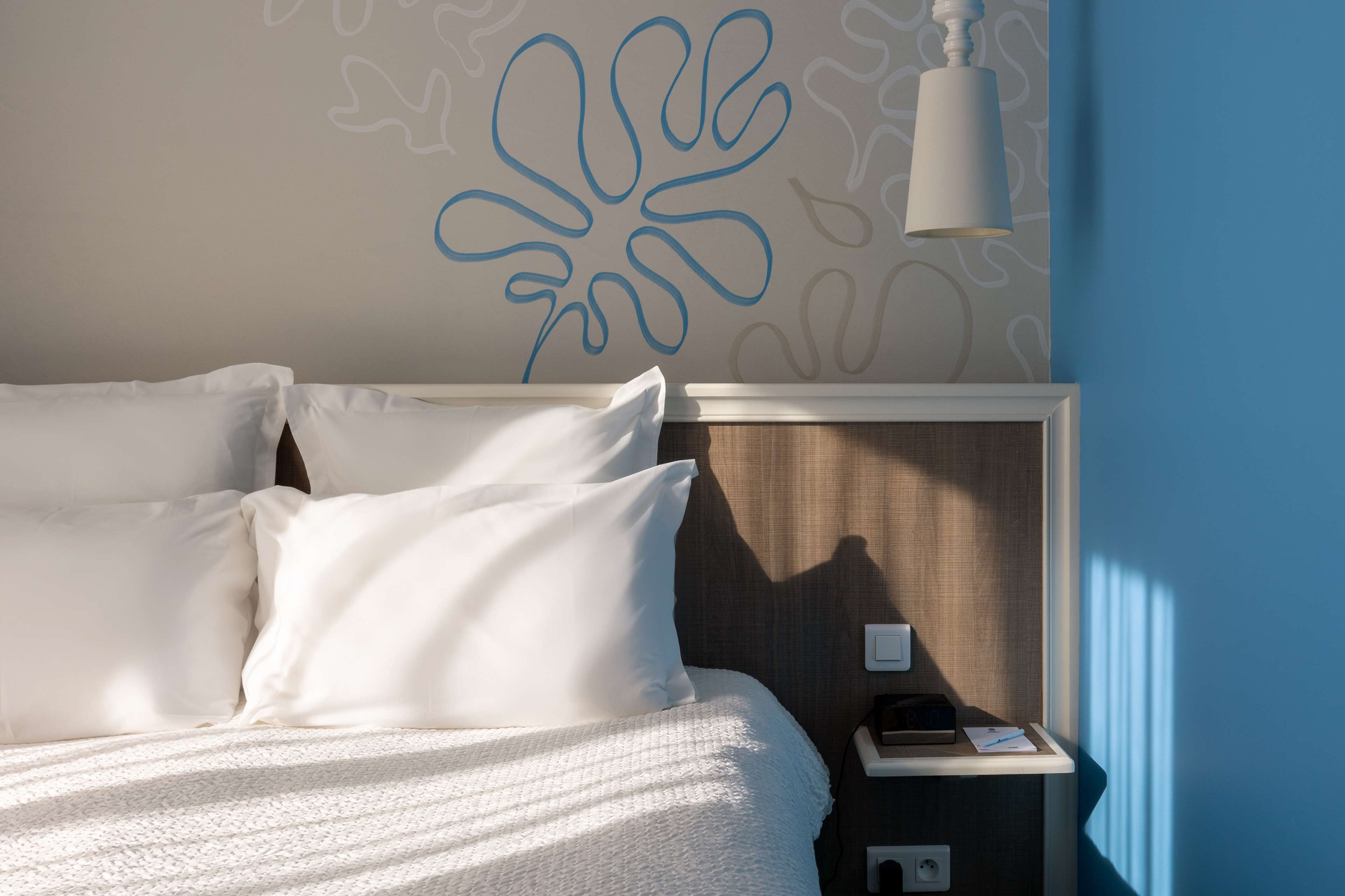A neatly made bed with white linens and pillows, a bedside table with a lamp, and a wall with abstract blue patterns.