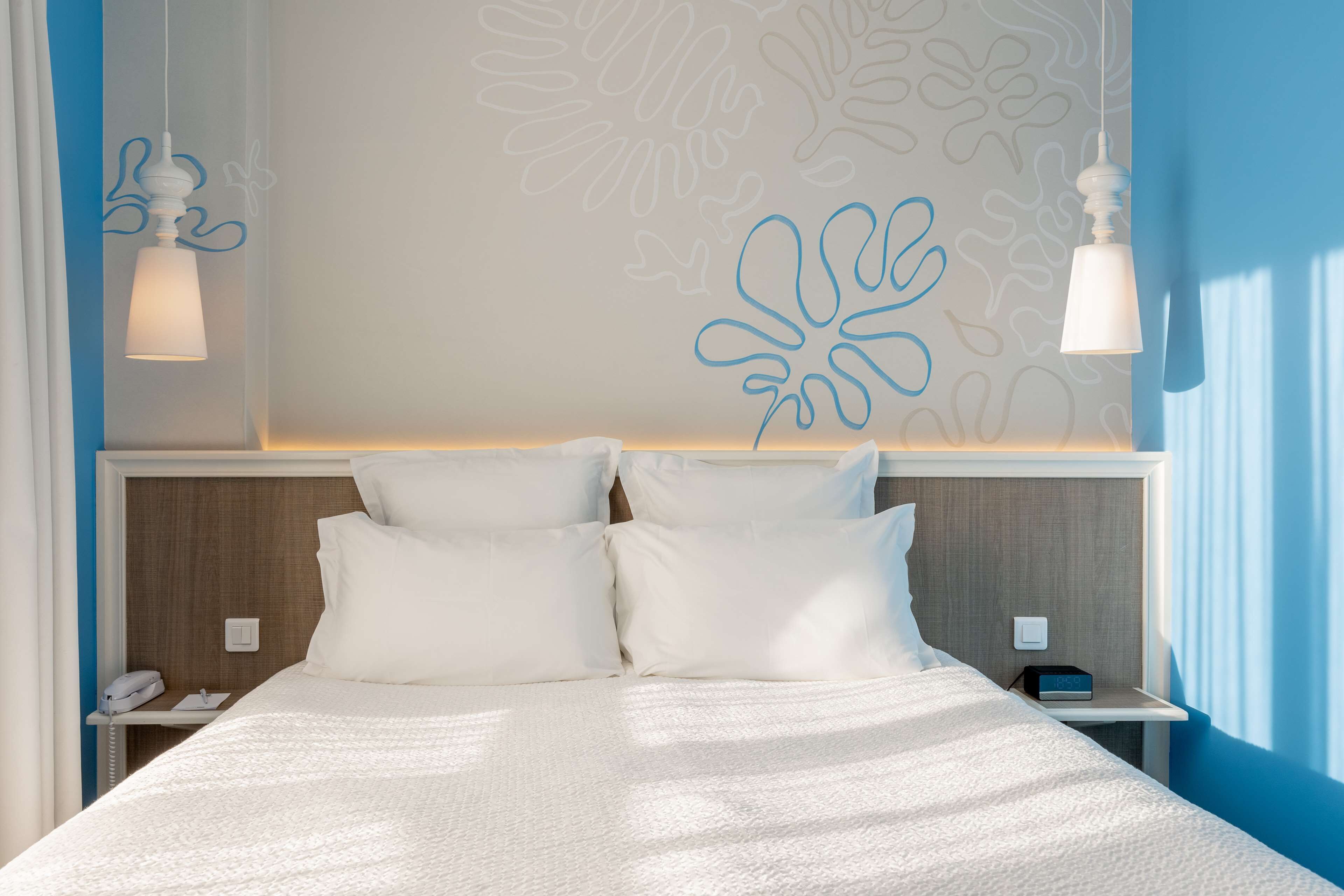 Una cama bien hecha con ropa blanca, una mesita de noche con un teléfono y un objeto pequeño, y una pared con una lámpara decorativa.