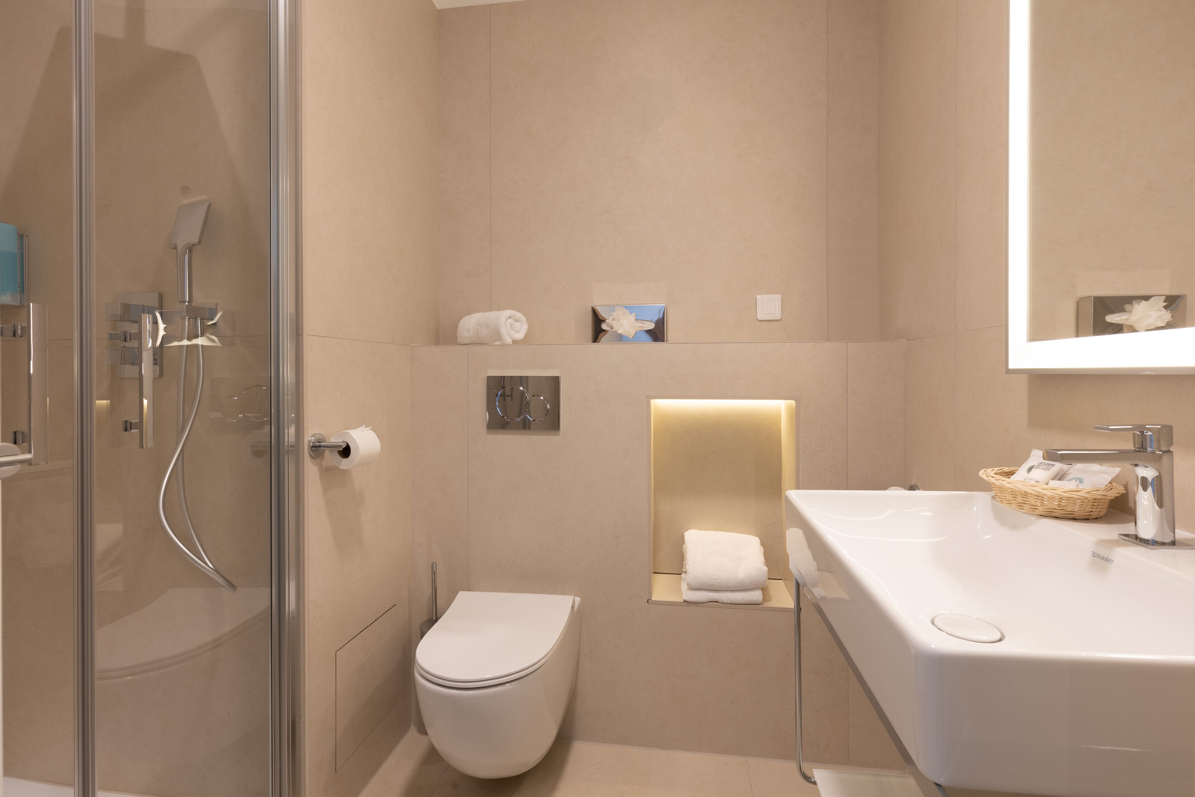Un baño moderno con ducha de vidrio, inodoro blanco, lavabo con una cesta de artículos de aseo y espejo sobre el lavabo.