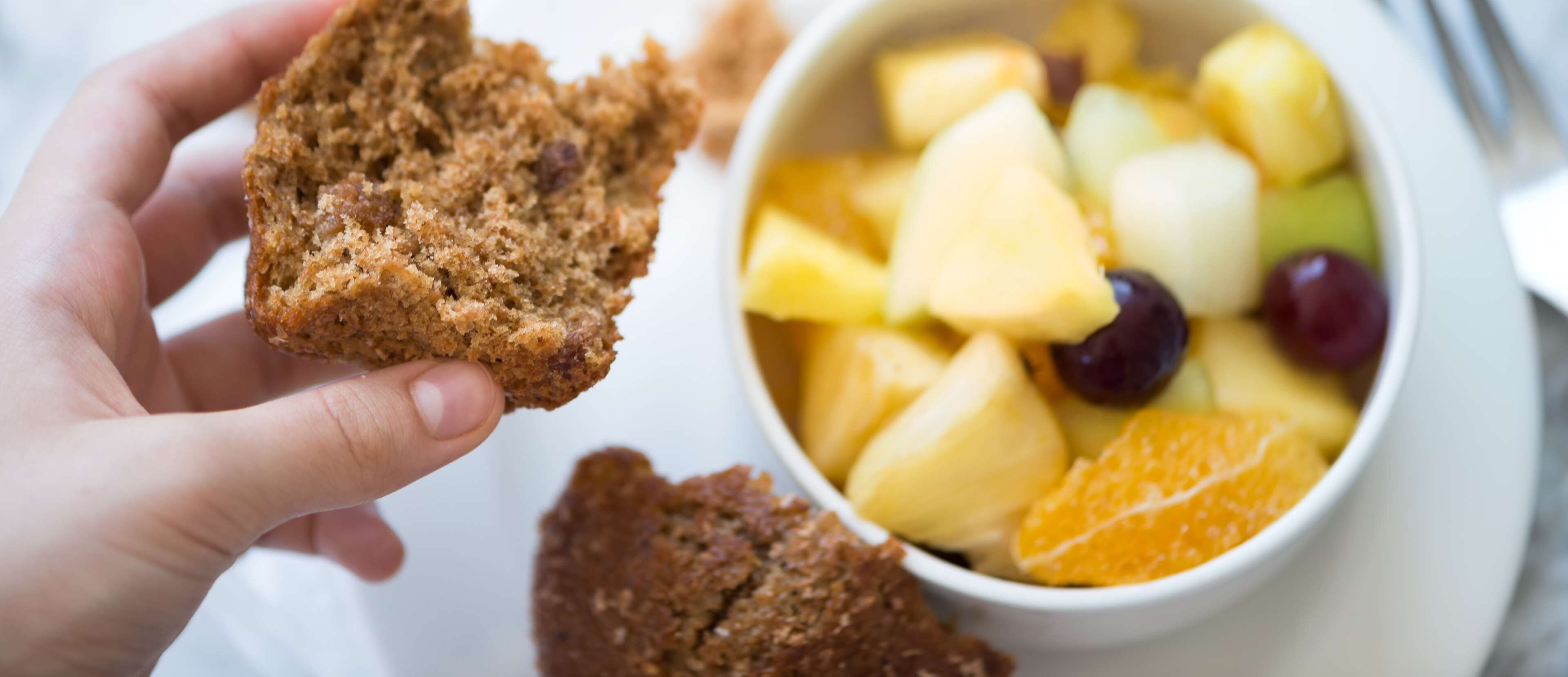 A hand holding a muffin next to a bowl of fruit.