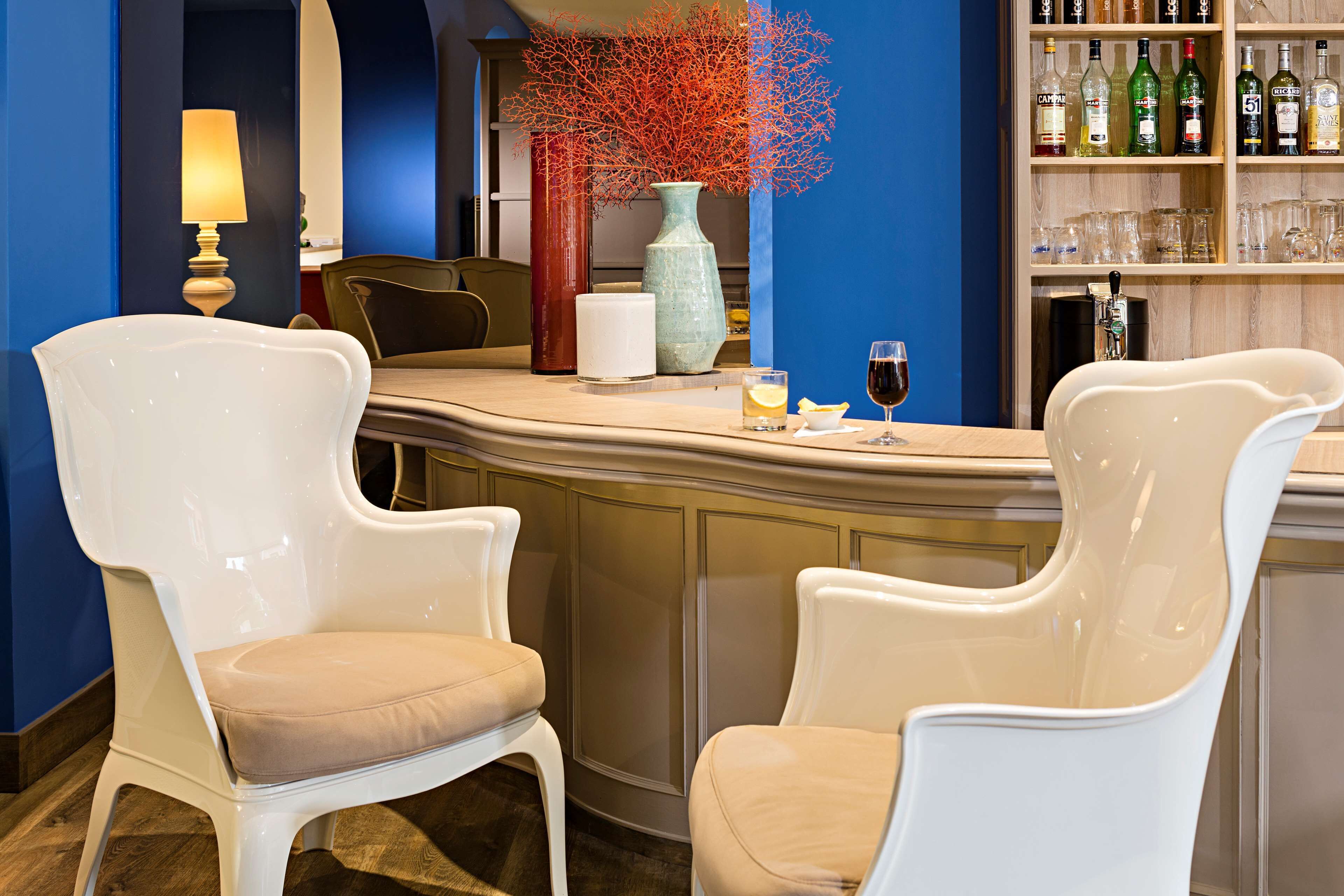 A bar area with two white chairs, a bar counter, a vase with red branches, and a shelf with bottles and glasses.
