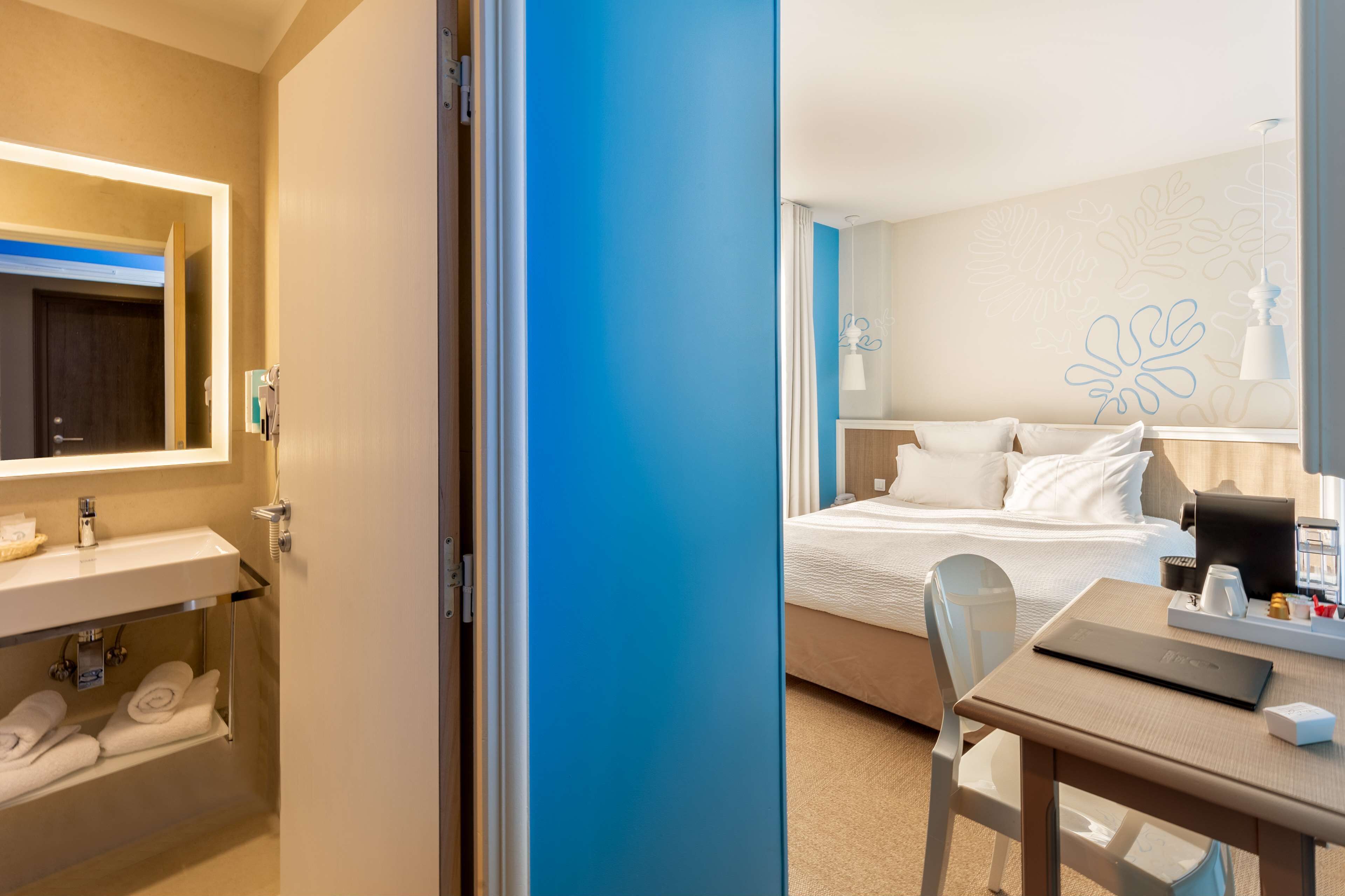 A hotel room with a bed, bedside table, and a desk with a laptop. There is a bathroom with a sink and towels.