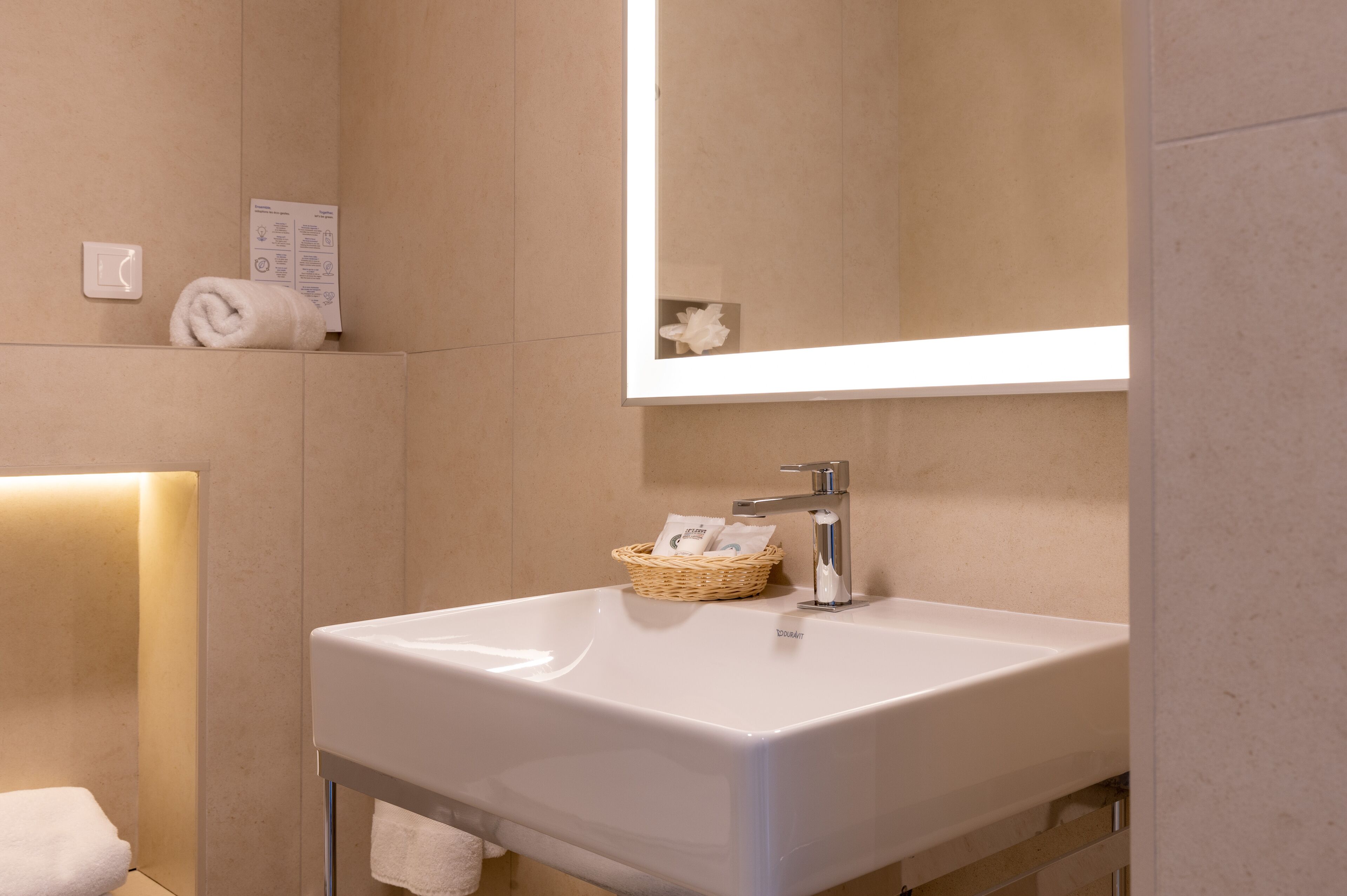 Un baño con lavamanos blanco, espejo, toalla, cesta con artículos de aseo e iluminación empotrada.