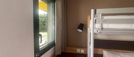 A compact hotel room with a bunk bed, a small wooden stool, a bedside lamp, and a window offering a view of greenery.