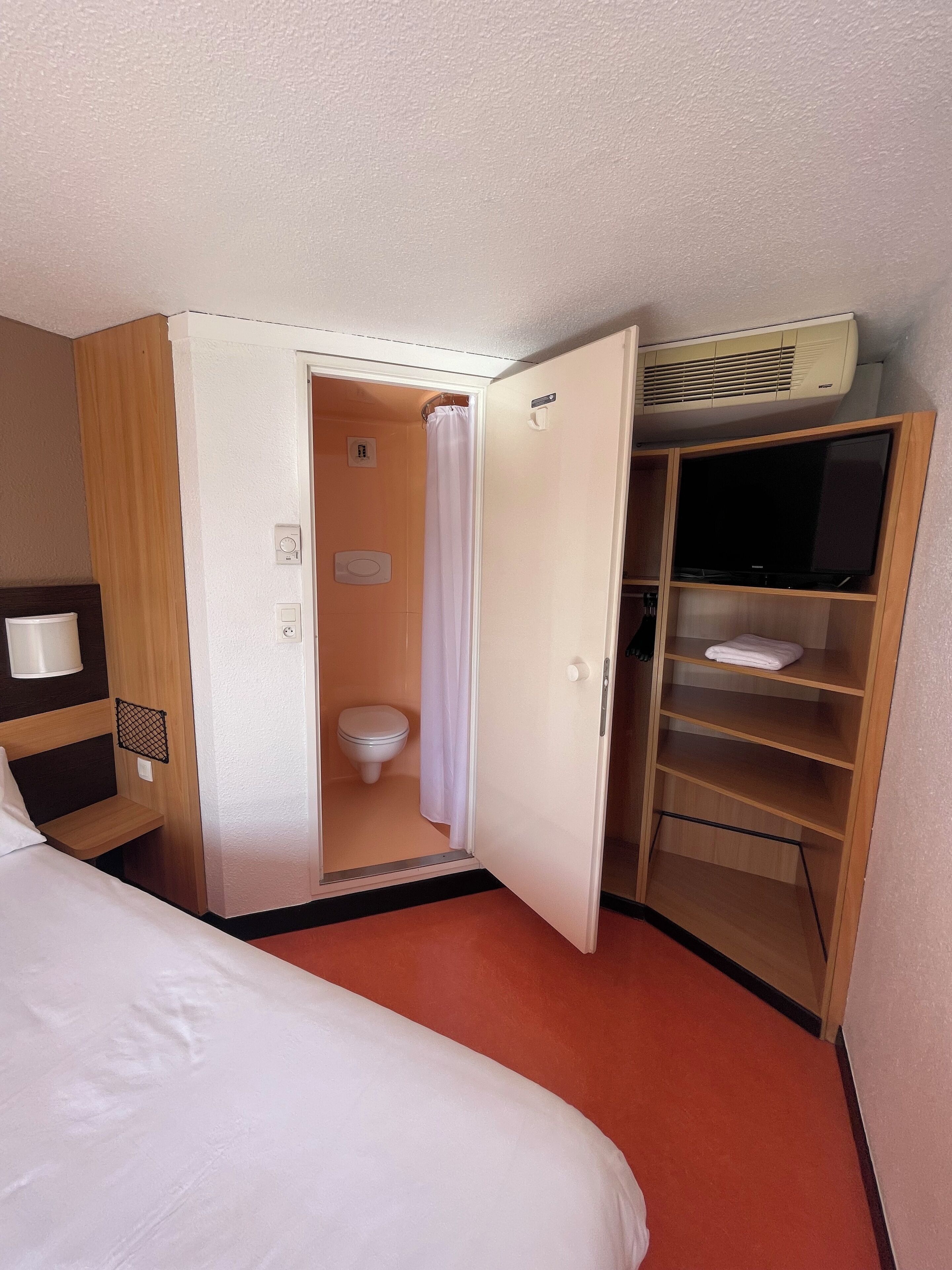 A hotel room with a bed, a bathroom with a toilet and shower, and a TV on a wooden shelf.