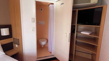 A hotel room with a bed, a bathroom with a toilet and shower, and a TV on a wooden shelf.