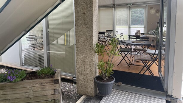 Een overdekt terras met tafels, stoelen, een plant in pot en een houten bank.