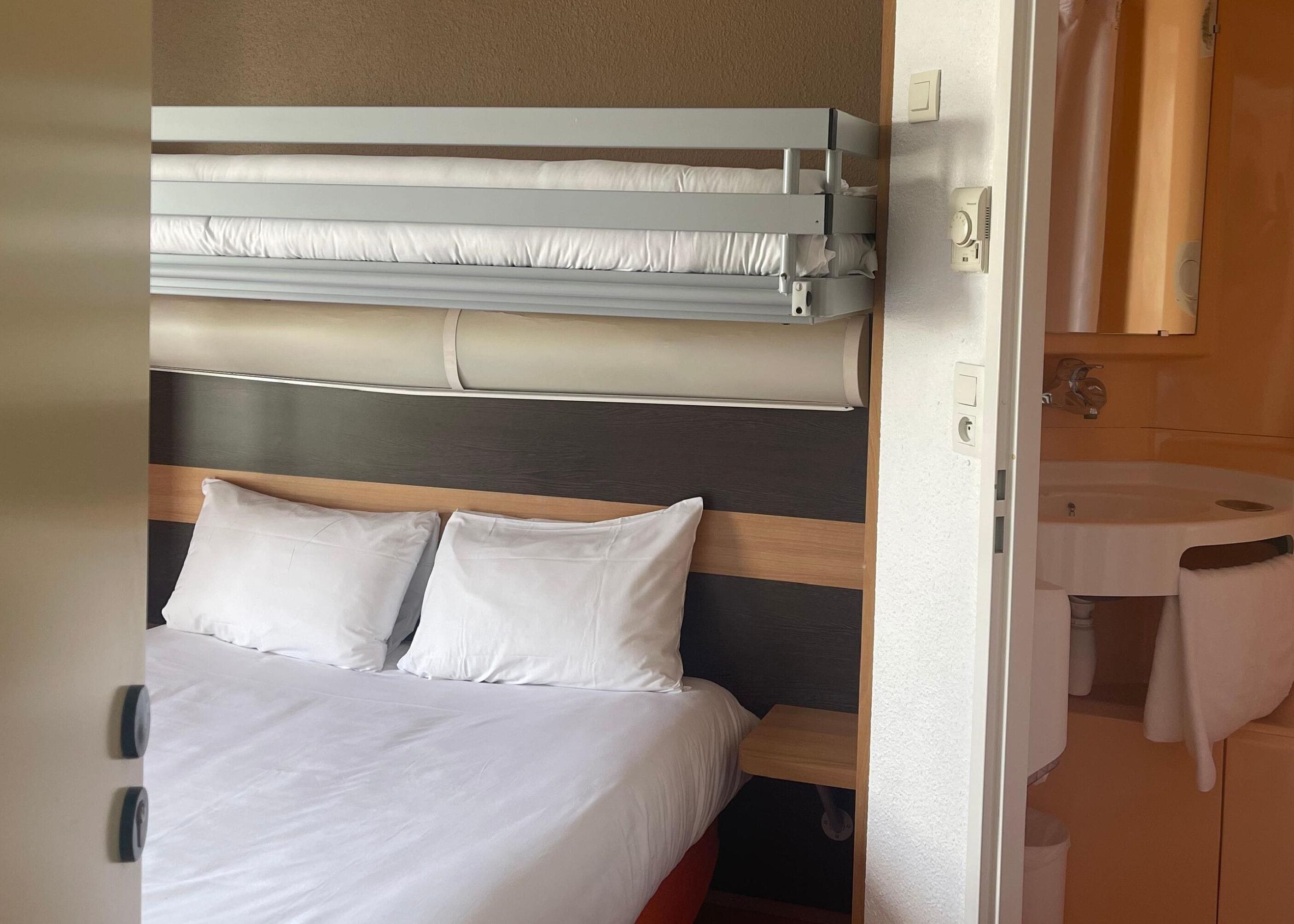 A bedroom with a bunk bed, a wooden headboard, a bedside table, and a bathroom with a sink and towel rack.