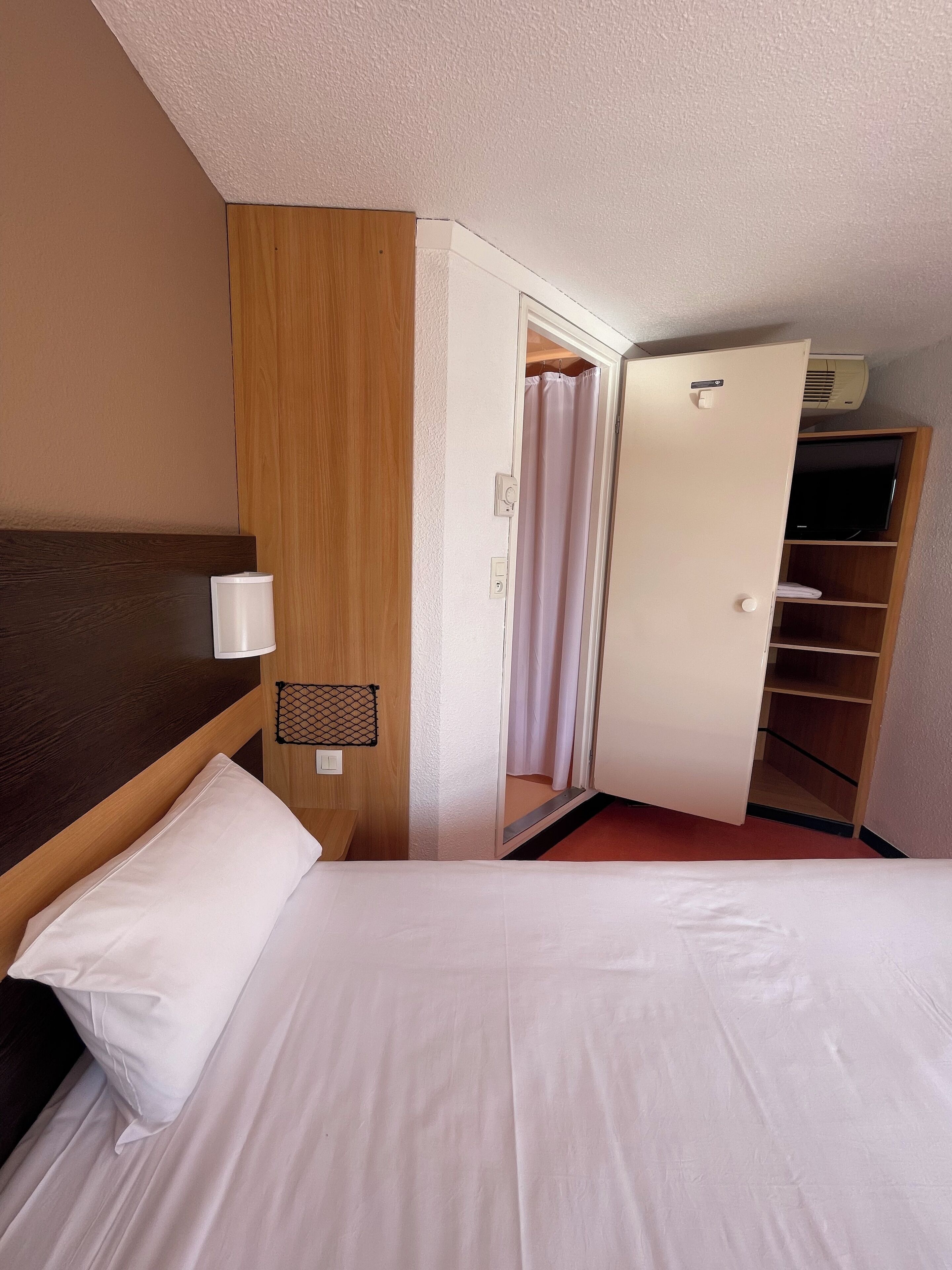 Hotellihuone, jossa on sänky, puinen vaatekaappi, televisio ja kattotuuletin.