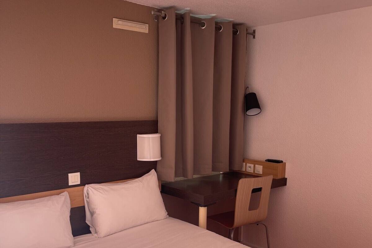 Habitació d'hotel amb llit, escriptori, cadira, làmpada i finestra amb cortines.