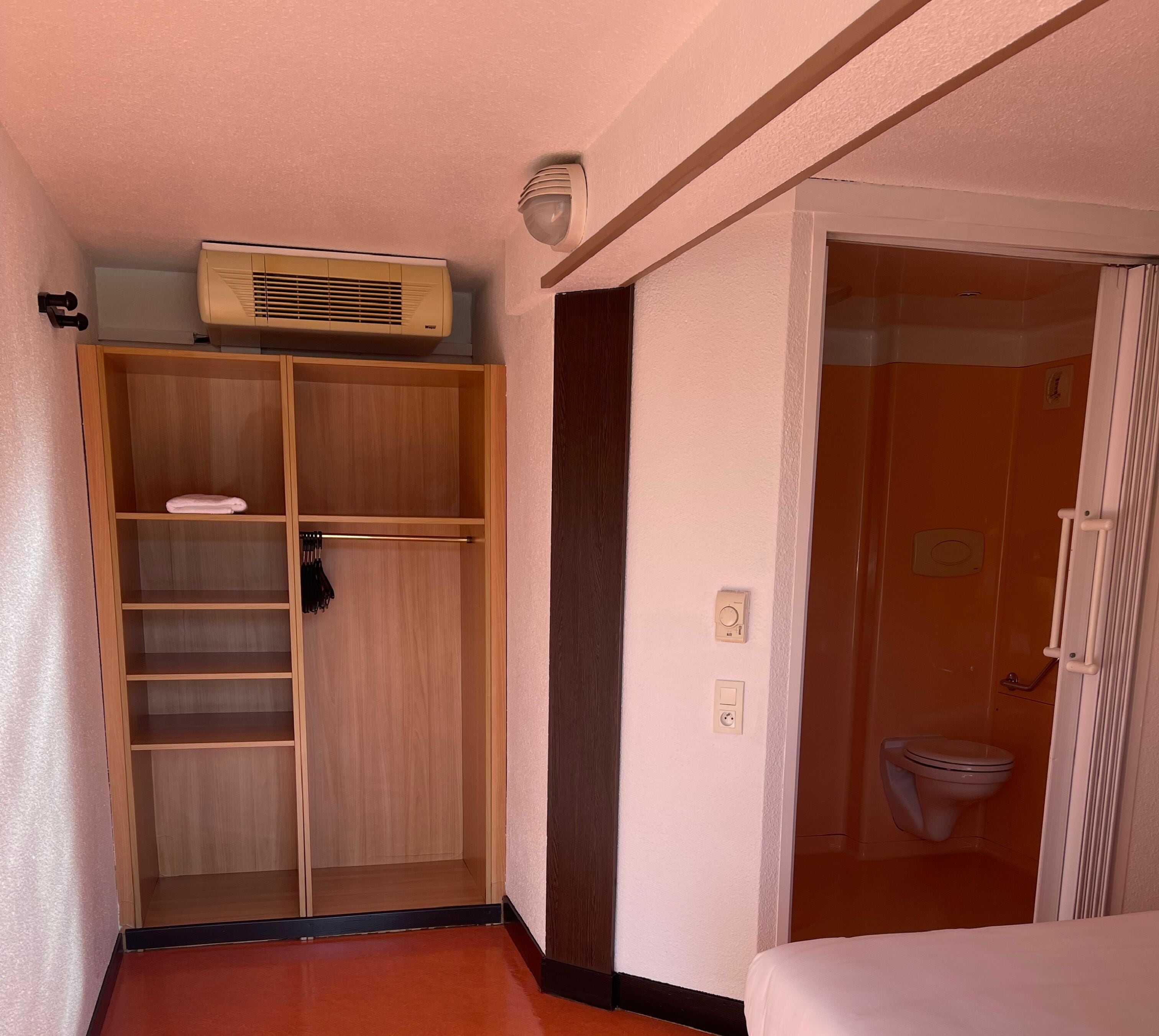 A hotel room with a wooden wardrobe, a ceiling fan, and a bathroom with a toilet and shower.