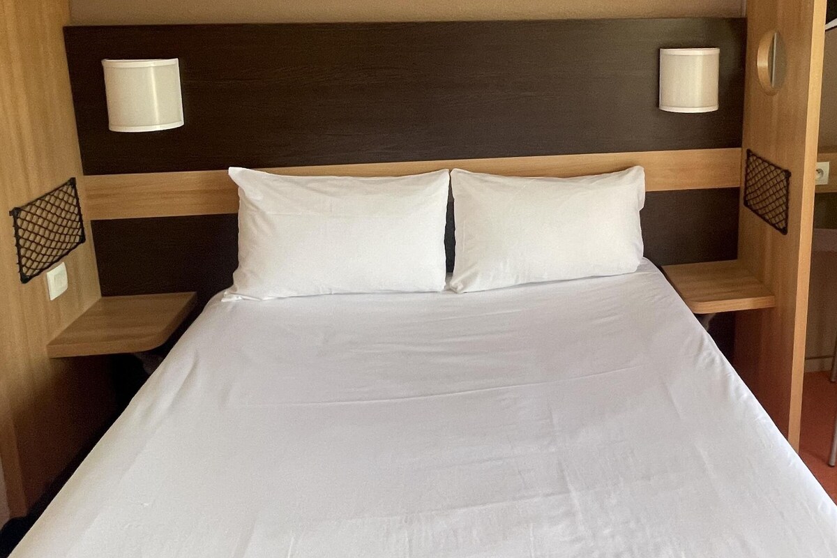 Una cama bien hecha con sábanas blancas, cabecera de madera y dos lámparas fijadas a la pared.