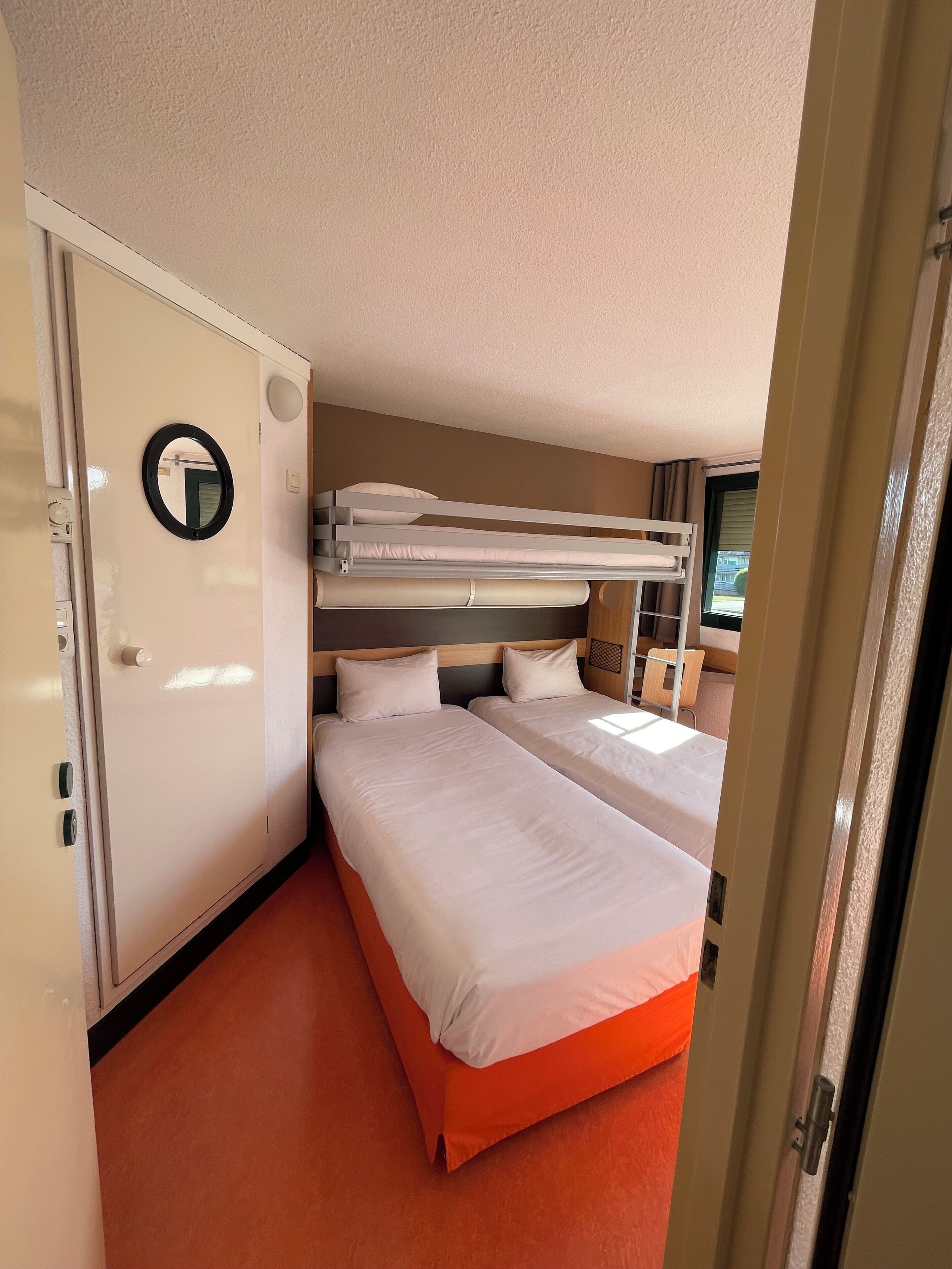 Quarto de hotel pequeno com uma cama beliche, uma cama de solteiro, um espelho e uma porta com puxador.