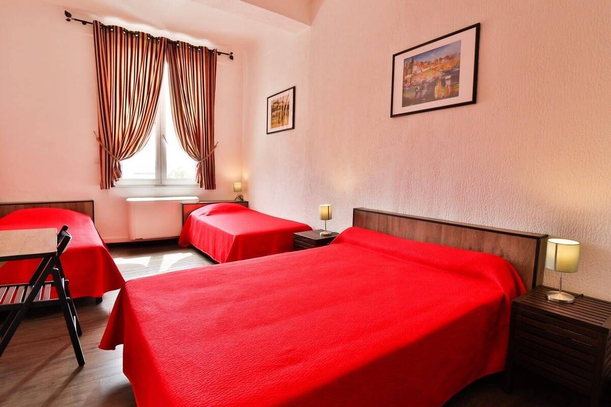 A hotel room with two single beds, a window with curtains, a bedside table with a lamp, and a framed picture on the wall.