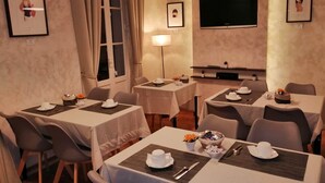 Un comedor con mesas dispuestas para cuatro, cada una con manteles blancos, cubiertos y tazas blancas. Se observan cuadros en las paredes, un televisor de pantalla plana y un amplio ventanal con cortinas.