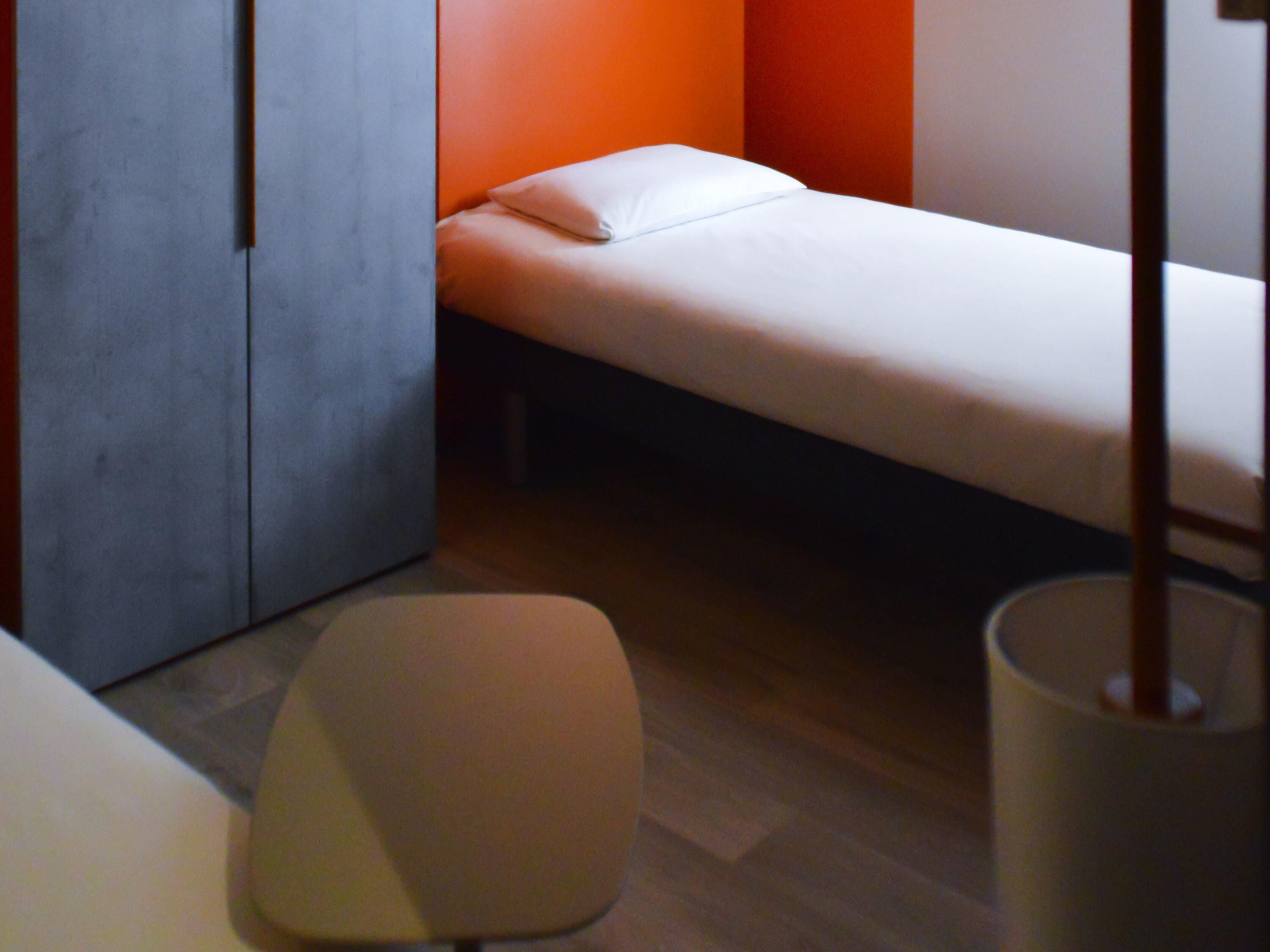 A small, single-bed room with a wooden floor, a white chair, and a bedside table.