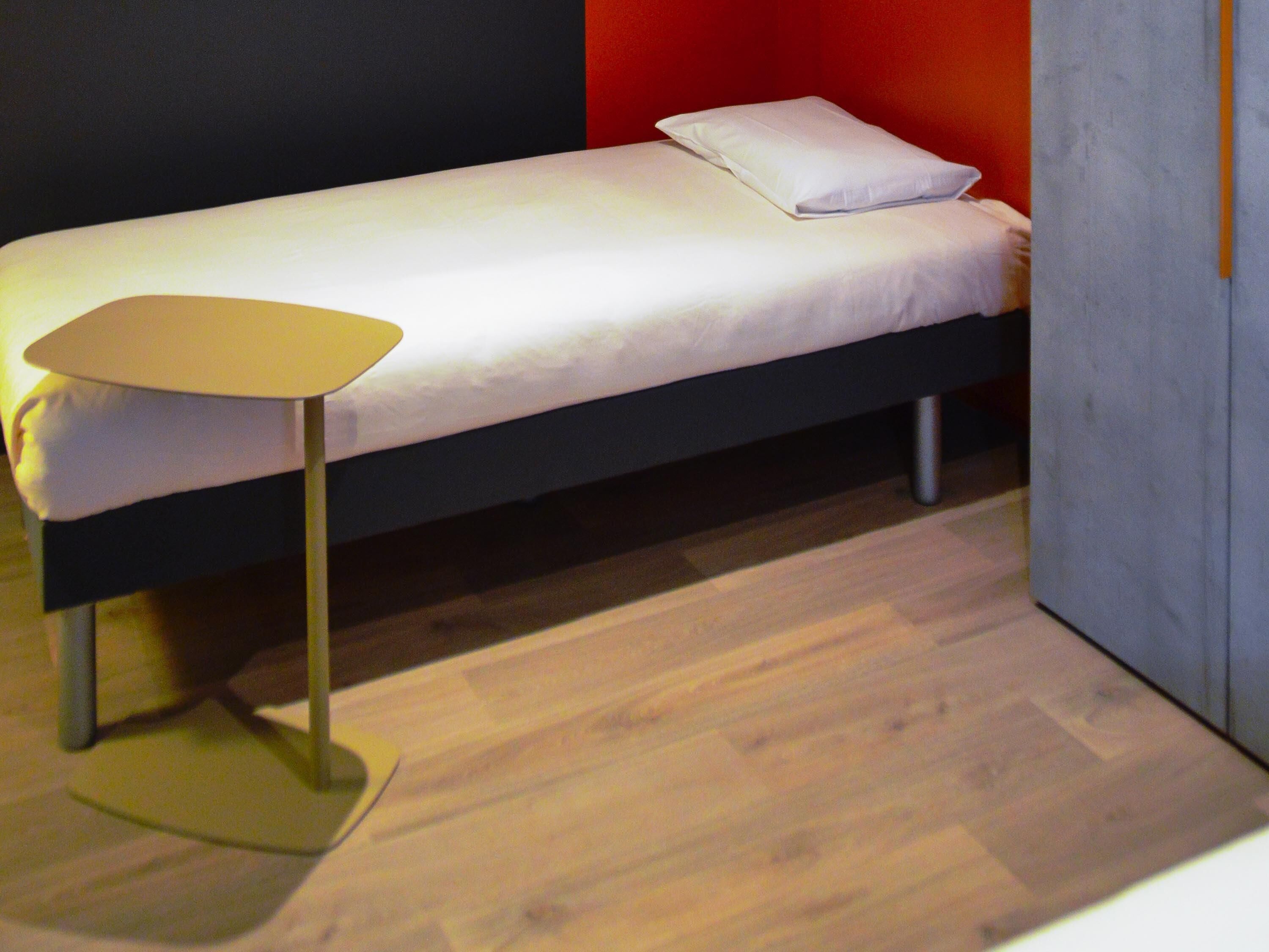Una cama individual con colchón y almohada blancos, una mesita con superficie amarilla y un suelo de madera.