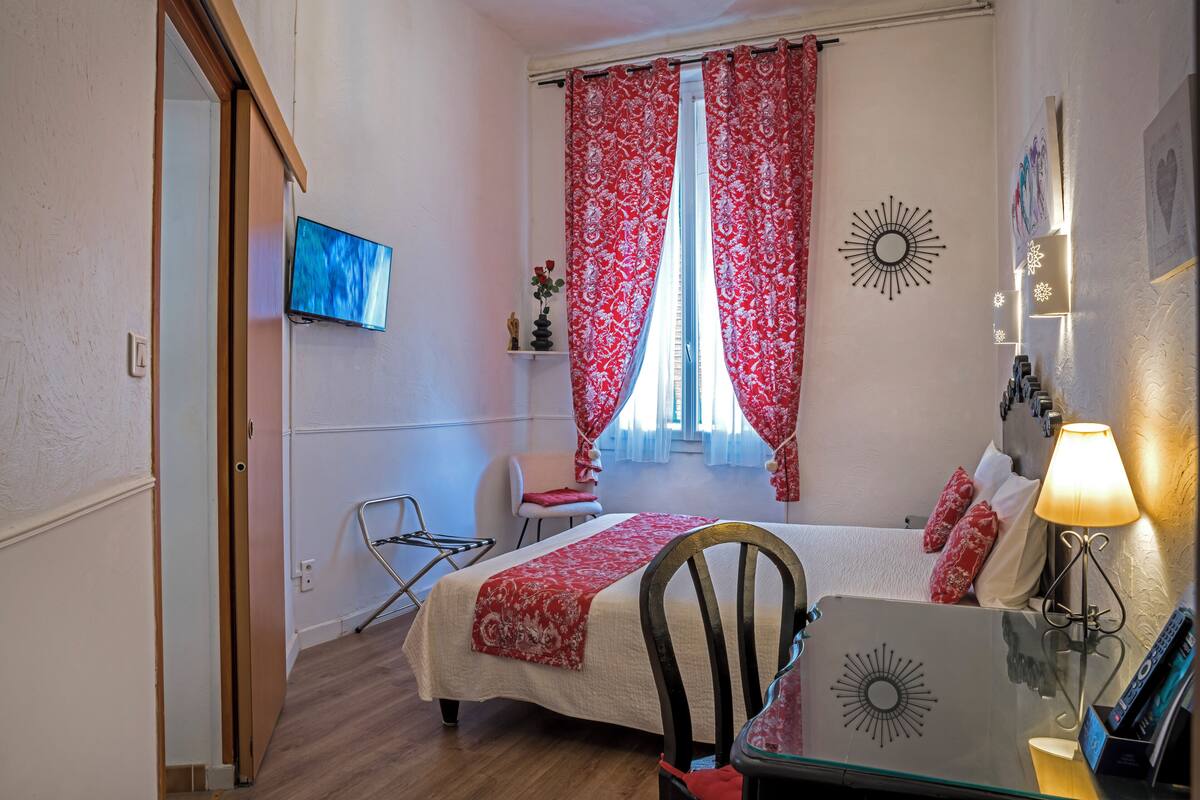 A hotel room with a bed, a desk with a lamp, a chair, a TV, and a window with red curtains.