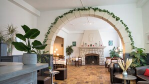 Un vestíbulo amplio con suelo de ladrillo, chimenea y un gran arco decorado con follaje. Hay varias mesas y sillas dispuestas para comer o conversar.