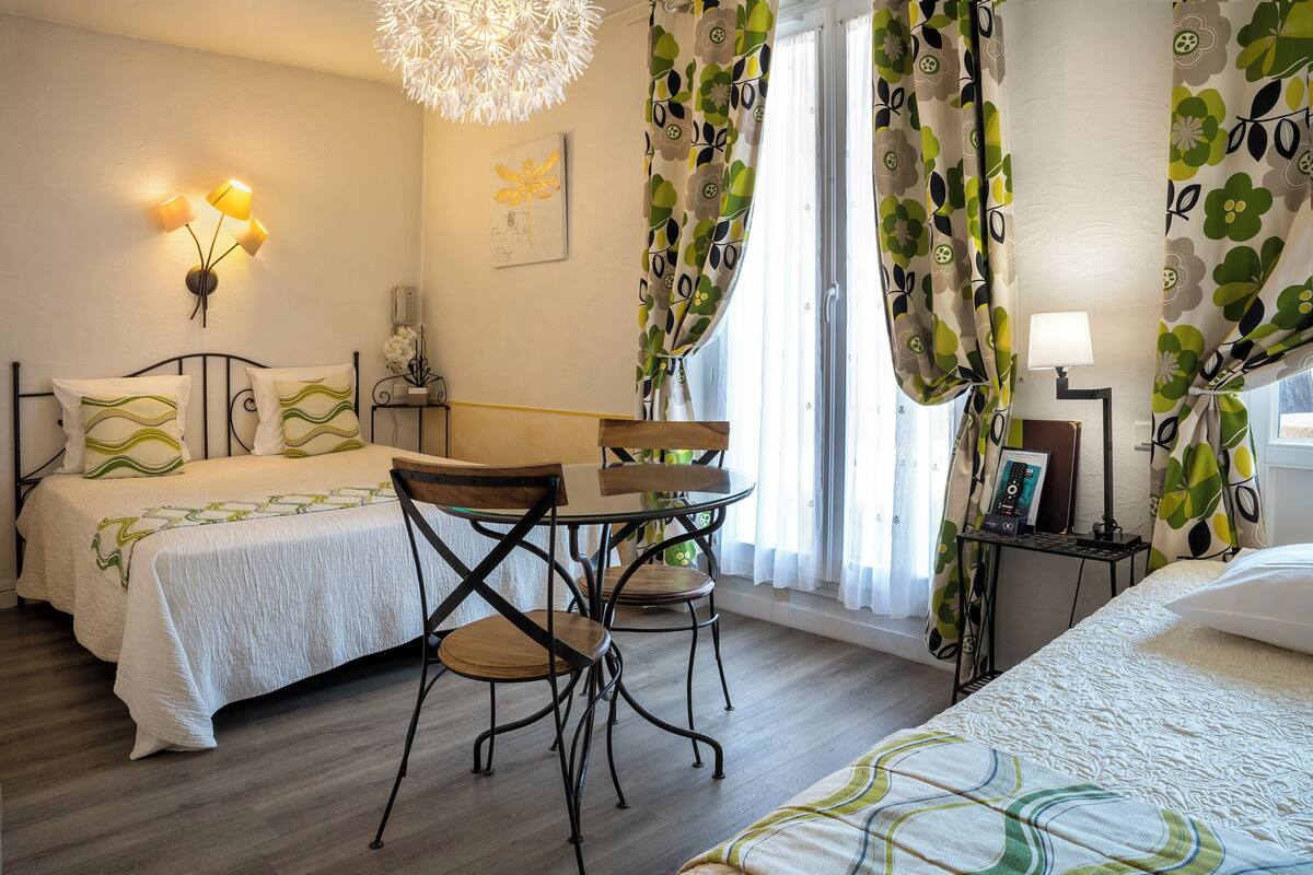 A hotel room with two beds, a dining table with chairs, a chandelier, and a bedside table with a lamp.