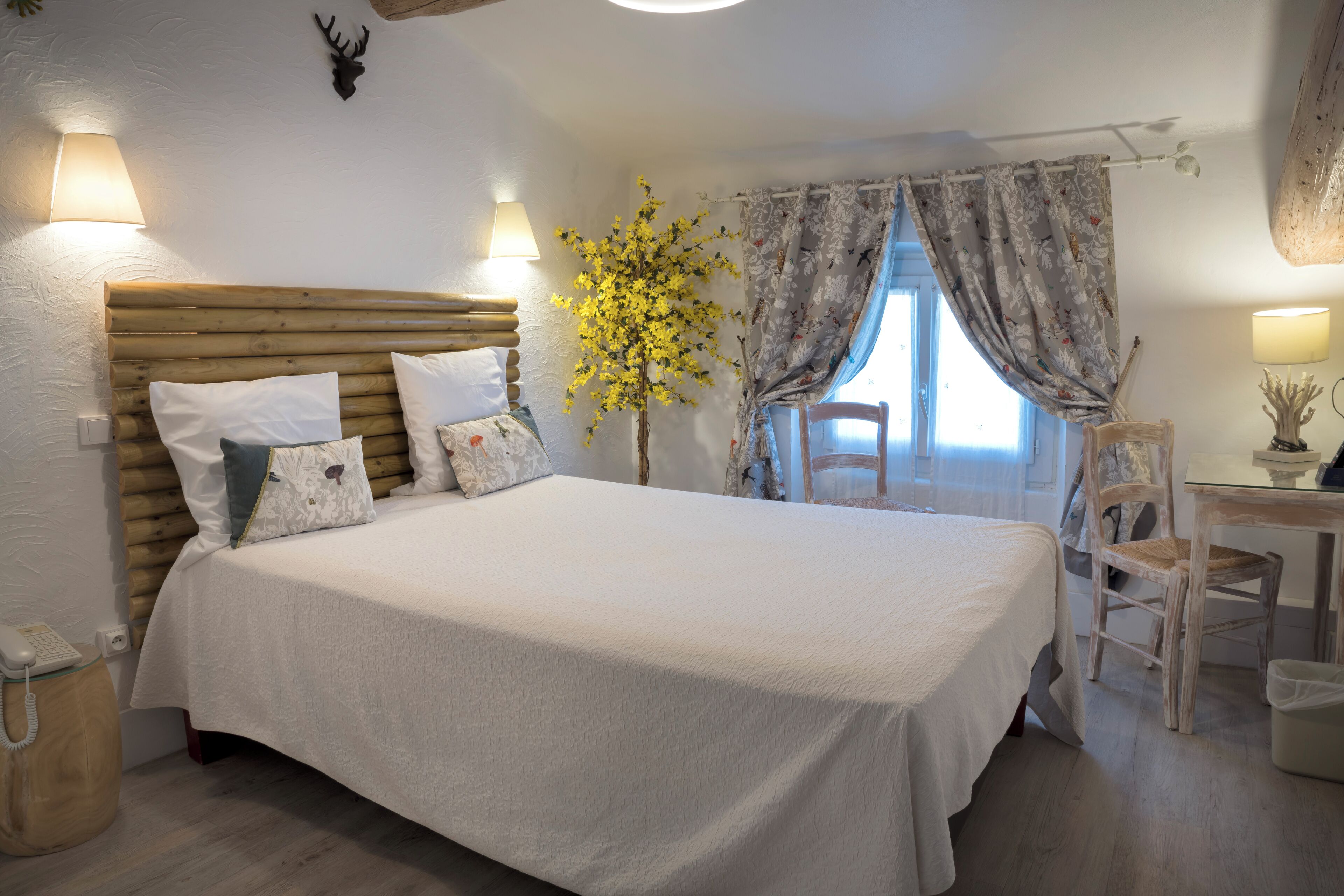 A bedroom with a wooden headboard, a bed with white bedding, a small table with a lamp, a chair, and a decorative plant.