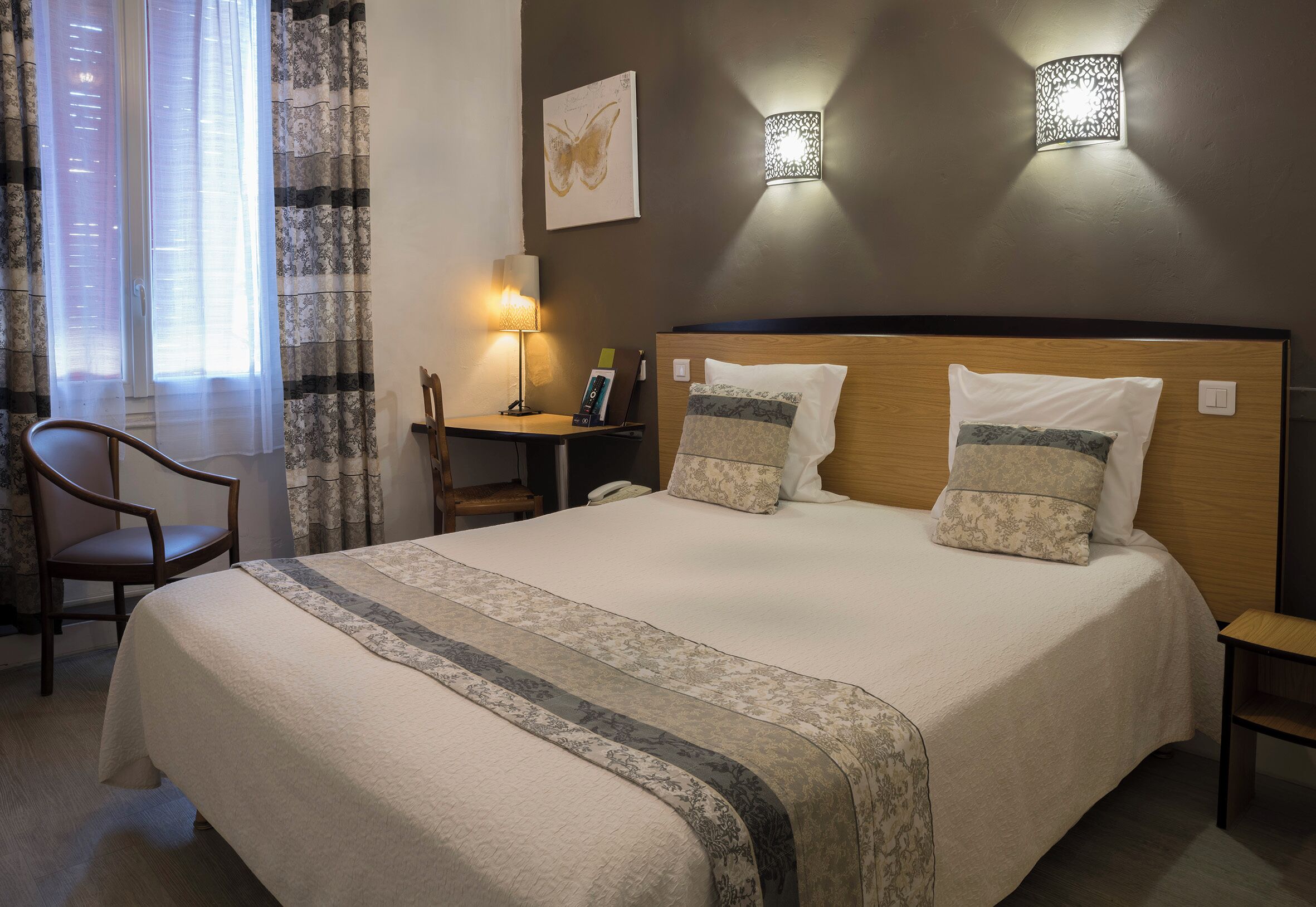 A neatly made bed with a headboard, a bedside table with a lamp, a chair, and a window with curtains.
