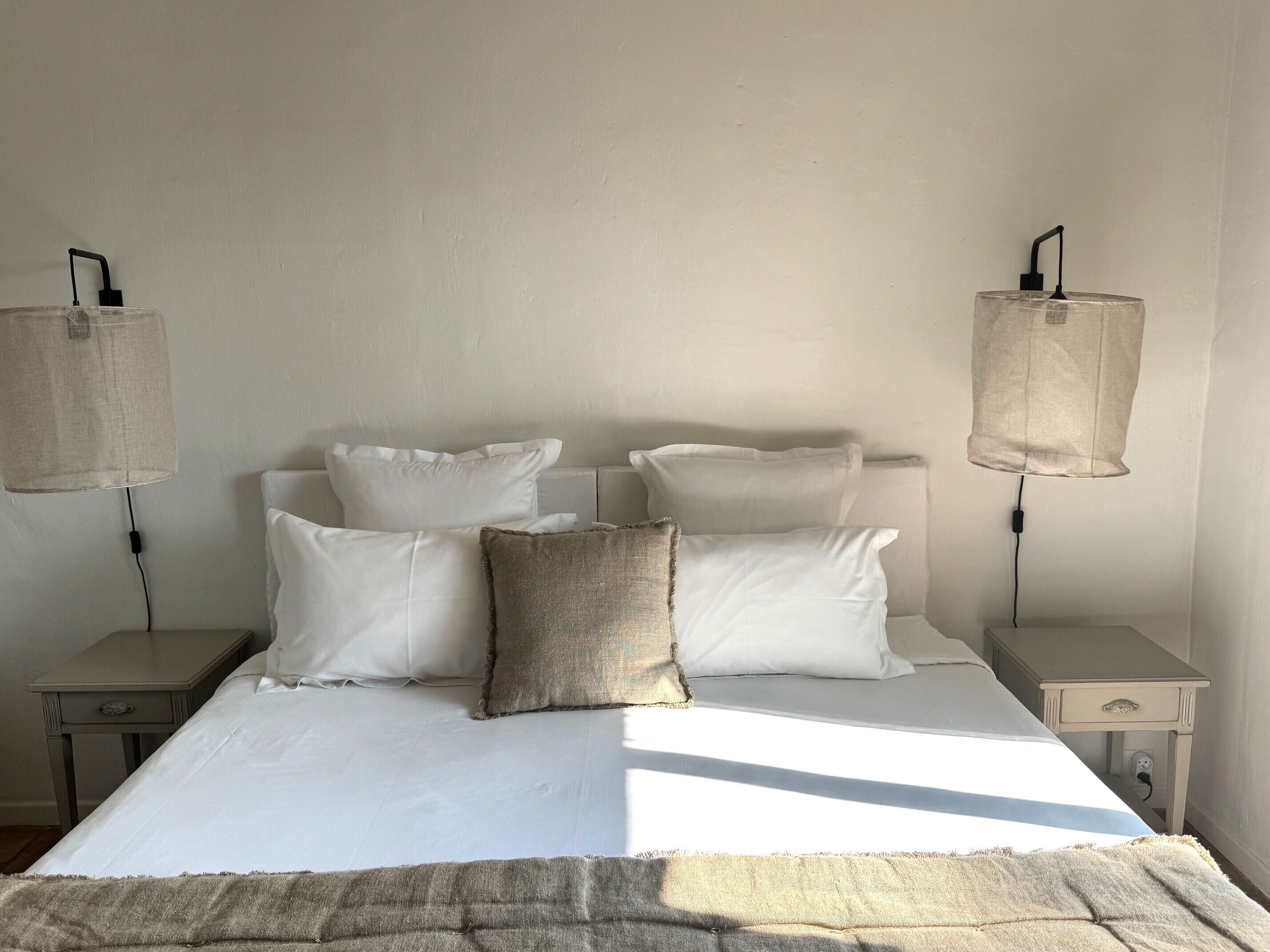 Un lit avec du linge de lit blanc et un plaid beige, deux tables de chevet avec des lampes, et un mur blanc uni.
