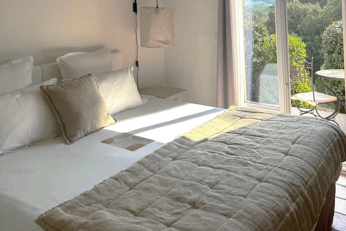 A bedroom with a large bed, a bedside table, a hanging lamp, and a view of the outdoors through a sliding glass door.