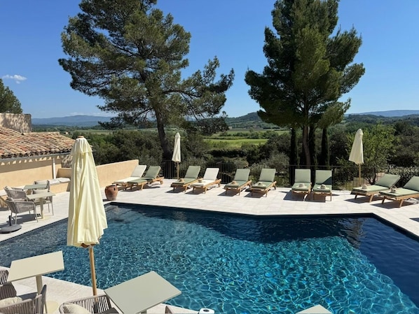 A poolside area with lounge chairs and umbrellas, surrounded by trees and a view of the countryside.