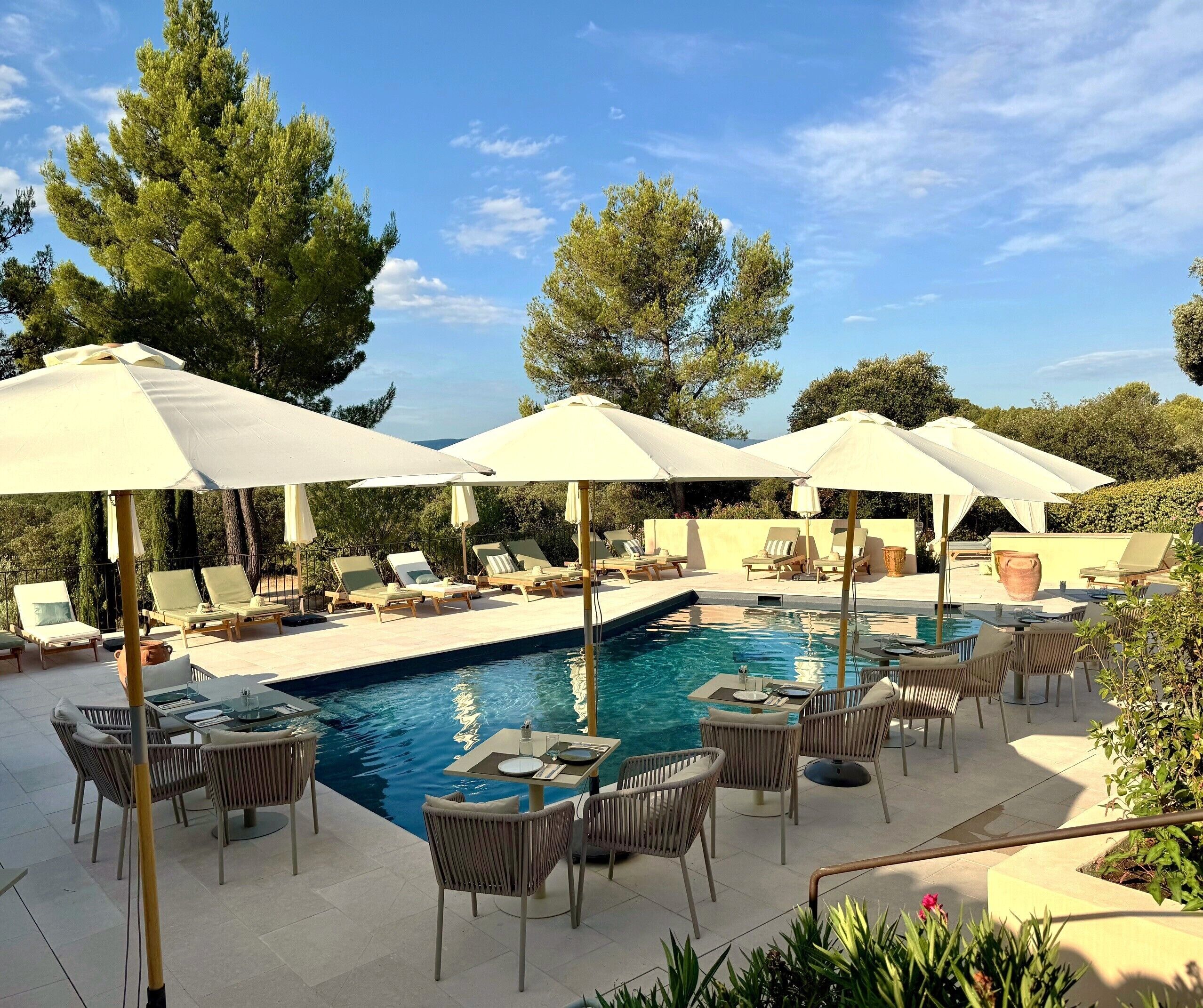 A poolside area with lounge chairs and umbrellas, surrounded by trees and a clear sky.