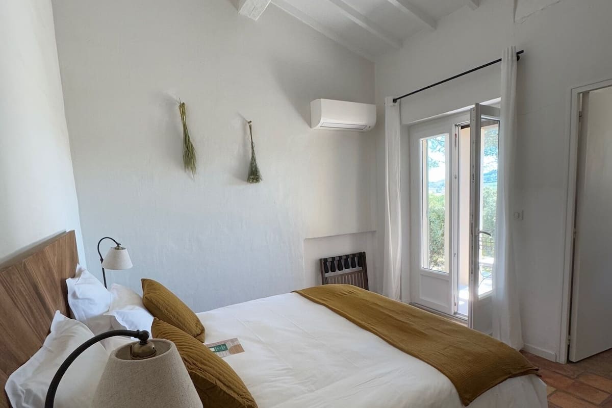 A bedroom with a large bed, white walls, a wooden headboard, a ceiling fan, a window with curtains, and a door leading outside.