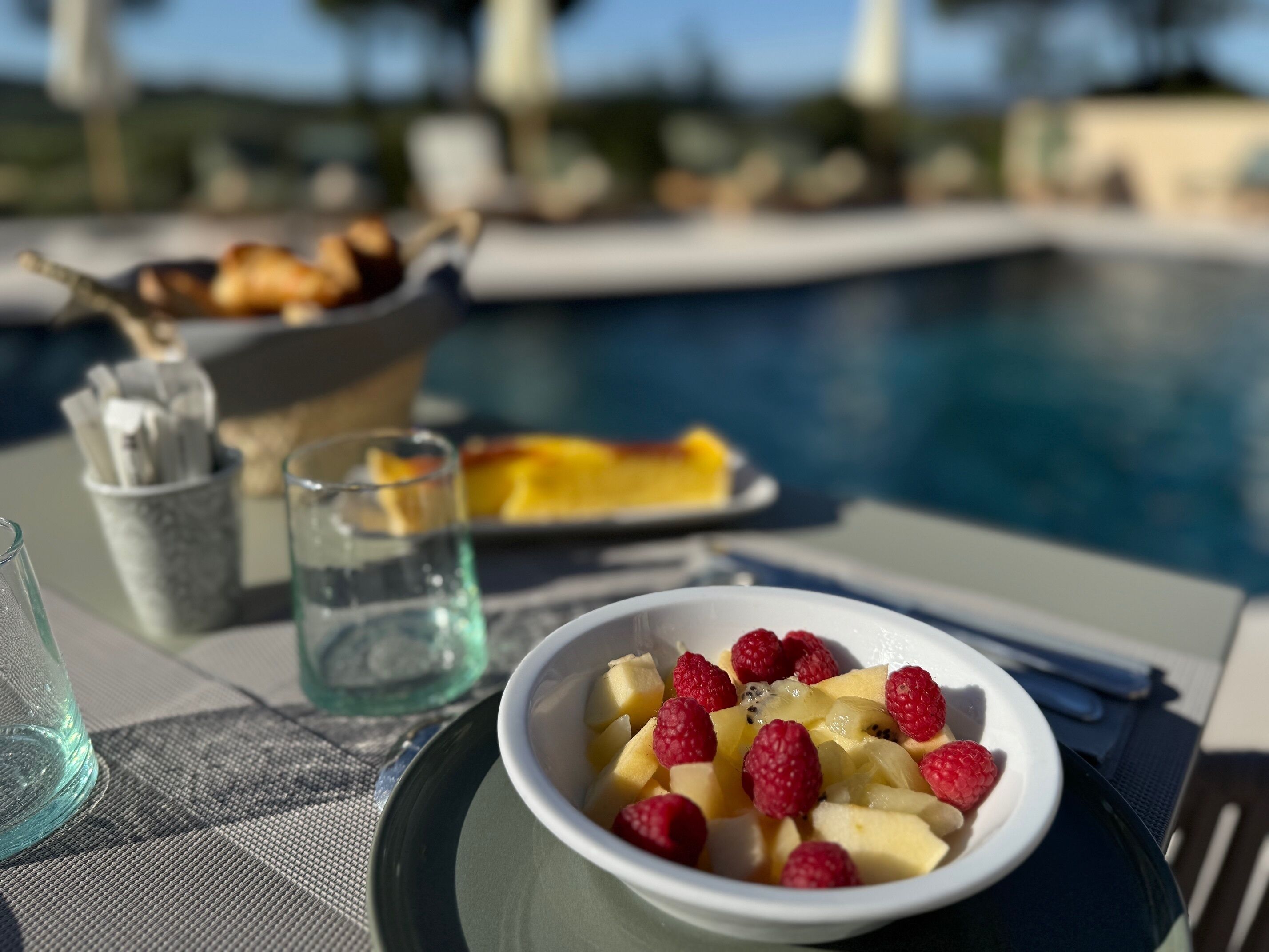 A table with a bowl of fruit salad, a glass of water, and a plate of food, near a poolside setting.