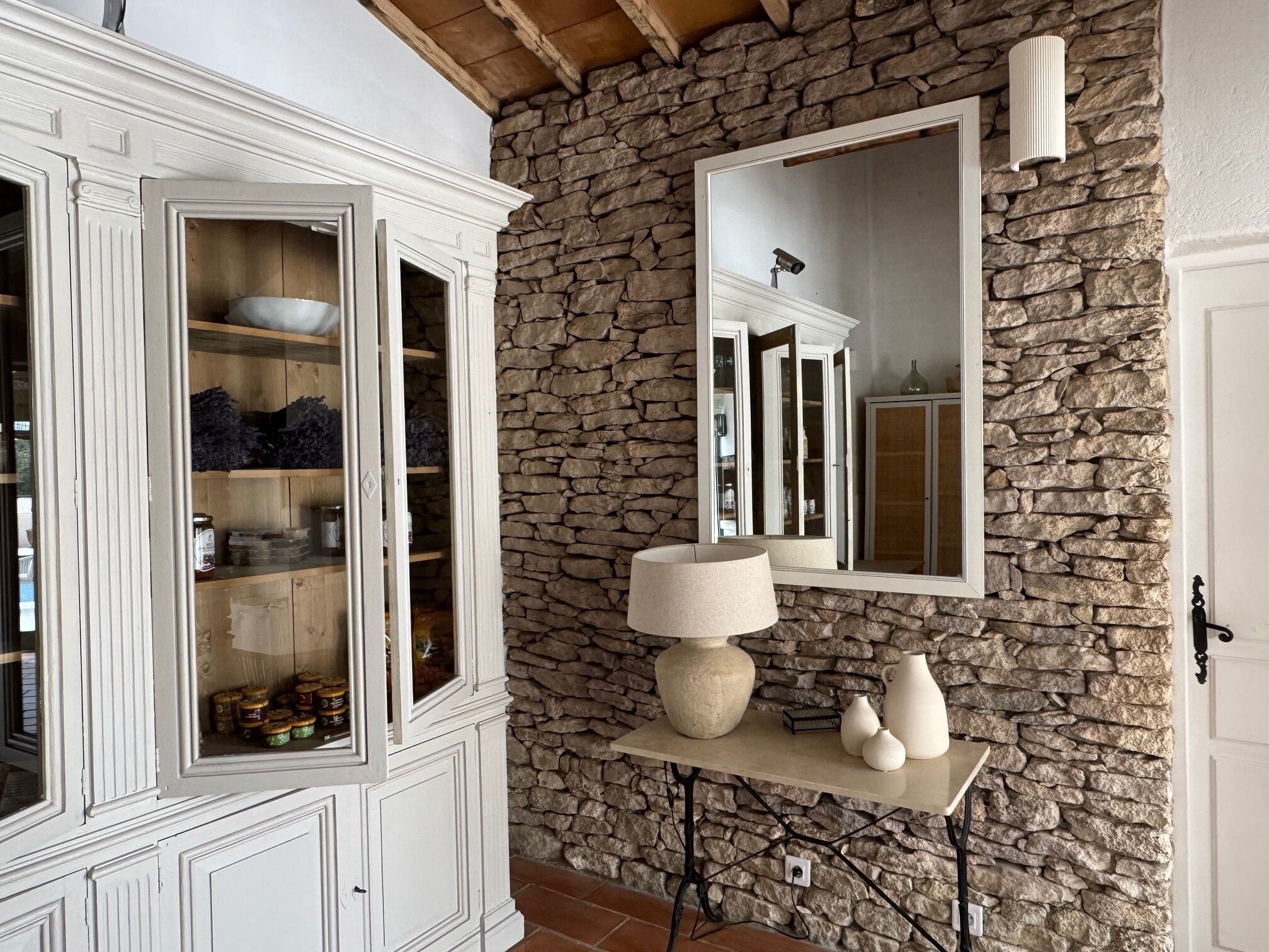 A room with a stone wall, a white cabinet with glass doors, a mirror, and a table with a lamp and vases.