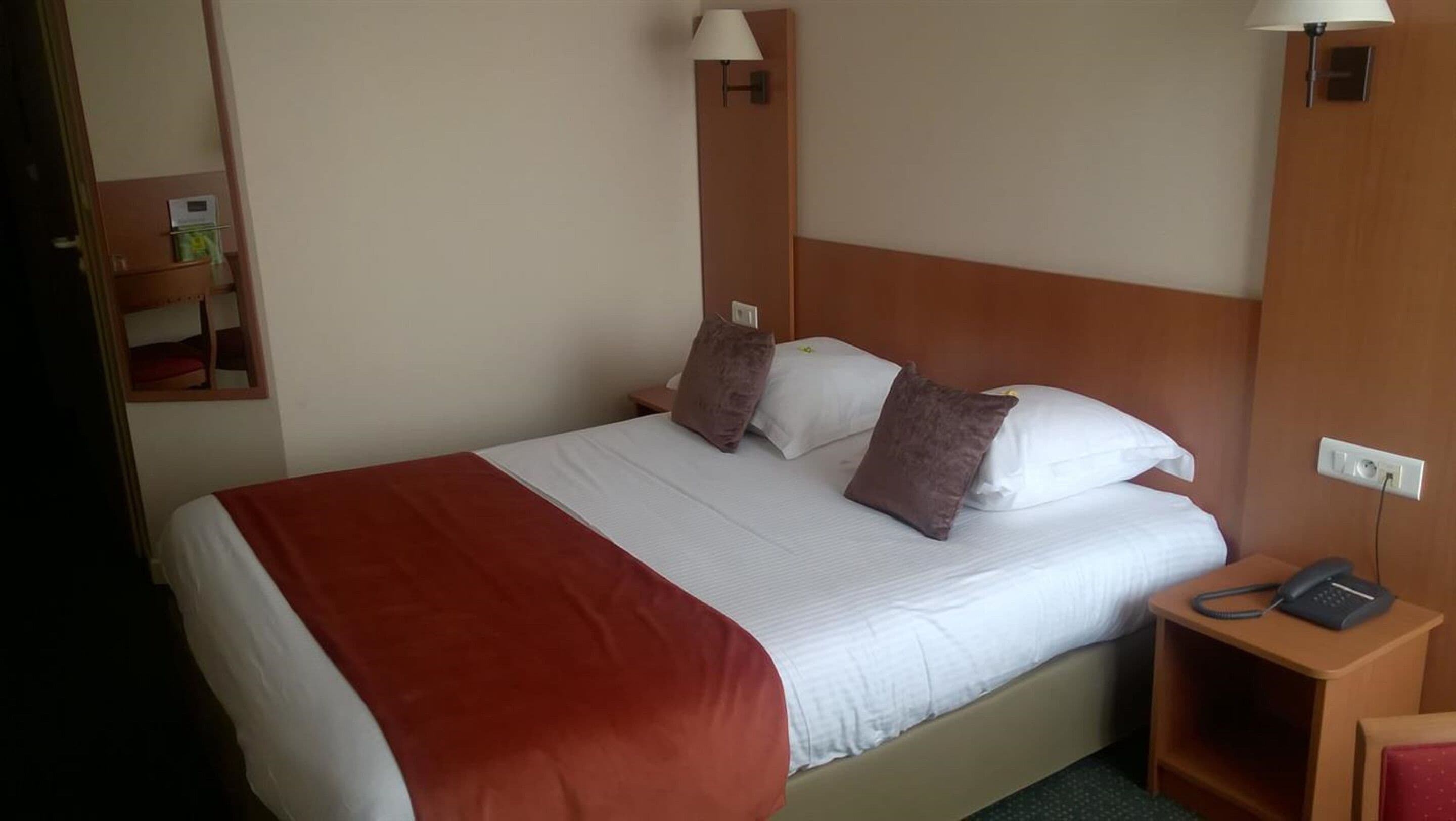 A hotel room with a bed, a bedside table with a phone, and a small shelf with a book.