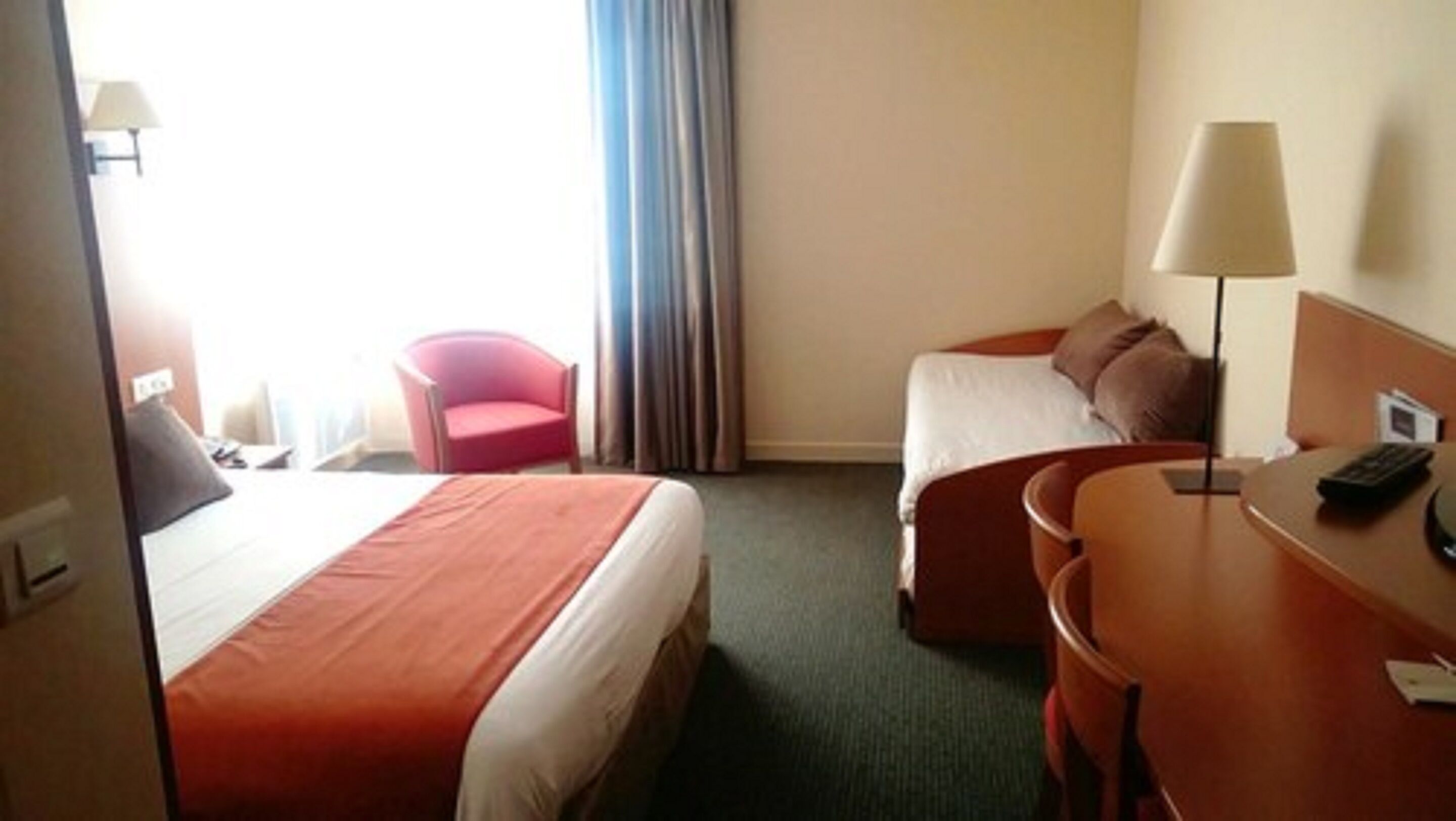 Une chambre d’hôtel avec deux lits, un bureau, une chaise, une lampe et une fenêtre avec des rideaux.