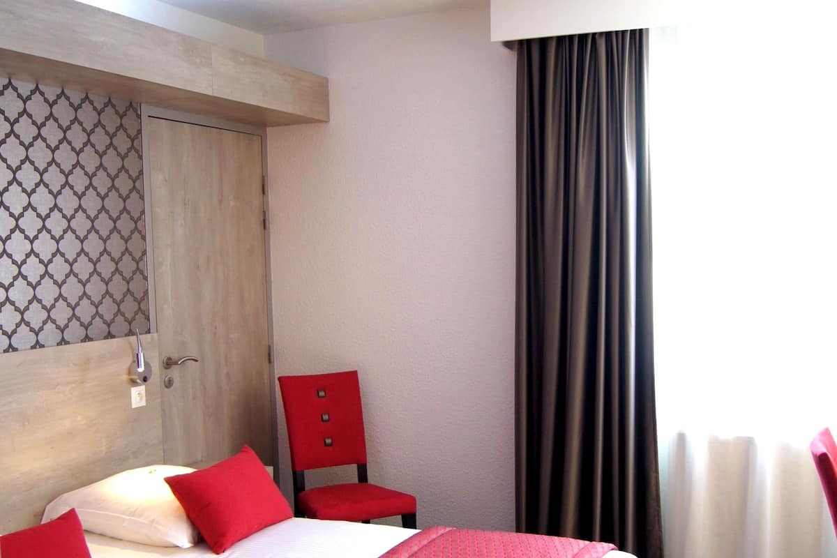 A hotel room with a large bed, red pillows, a wooden headboard, a red chair, and a window with curtains.