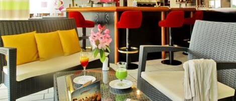 A bar area with a counter and stools, a sofa with yellow cushions, and a glass table with a magazine and drinks.