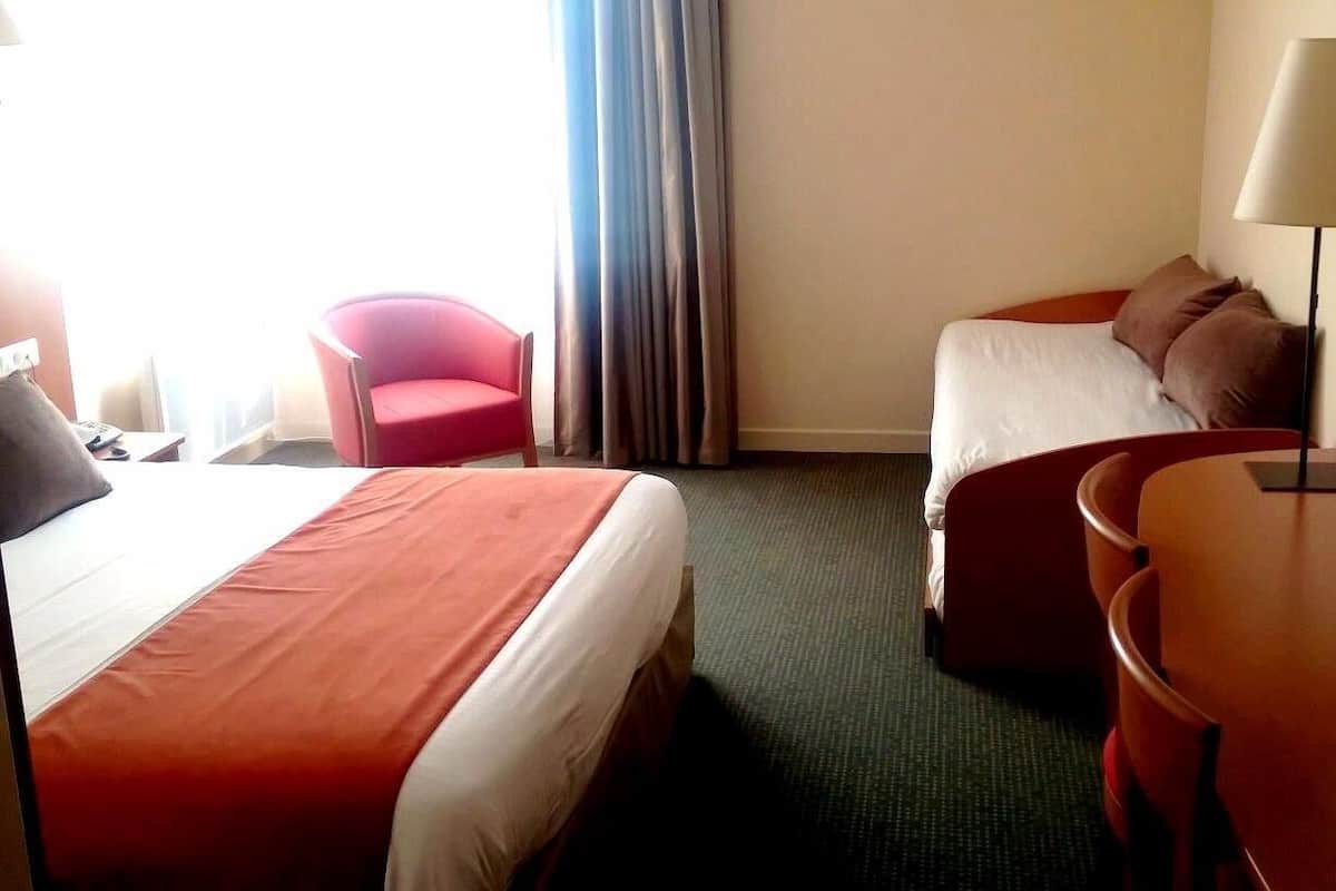 A hotel room with two beds, a desk, a chair, and a lamp.