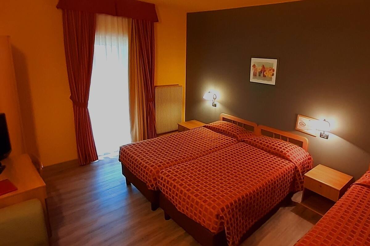 A hotel room with two beds, a nightstand, a lamp, a radiator, and a window with curtains.