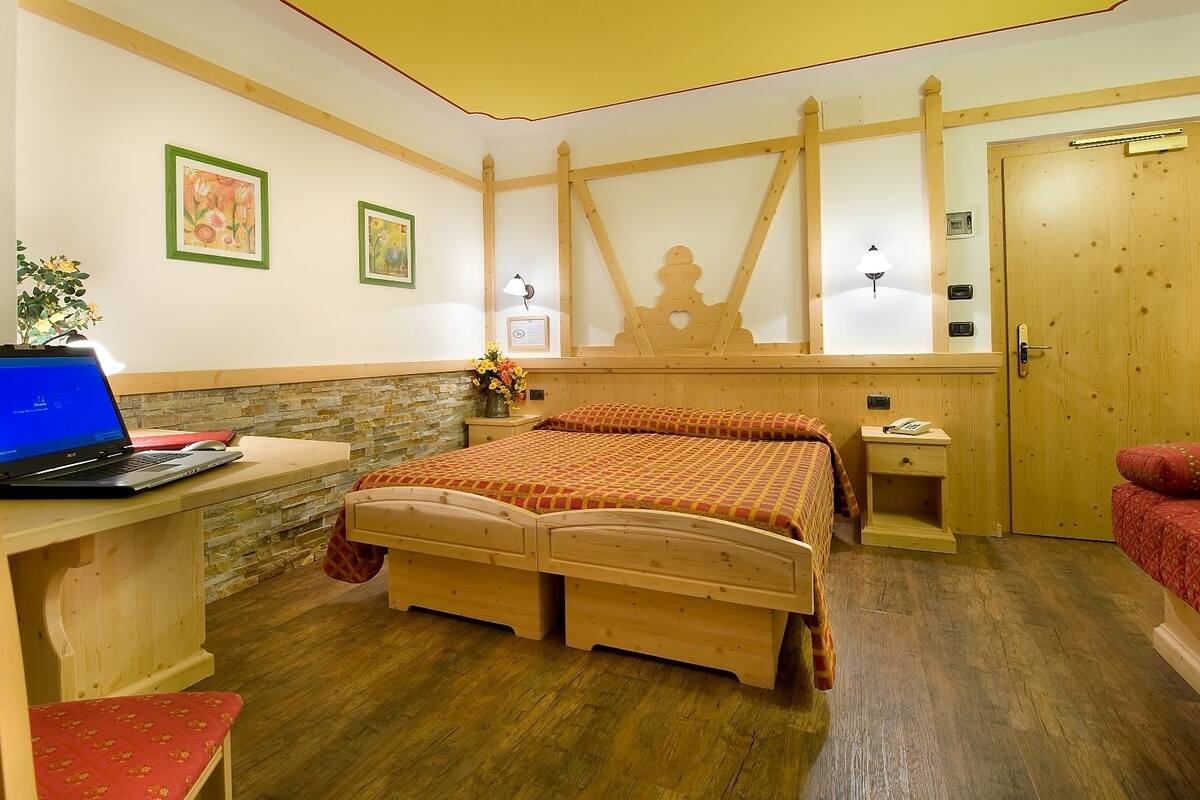 A room with a wooden bed, a desk with a laptop, a bedside table with a lamp, and a door.